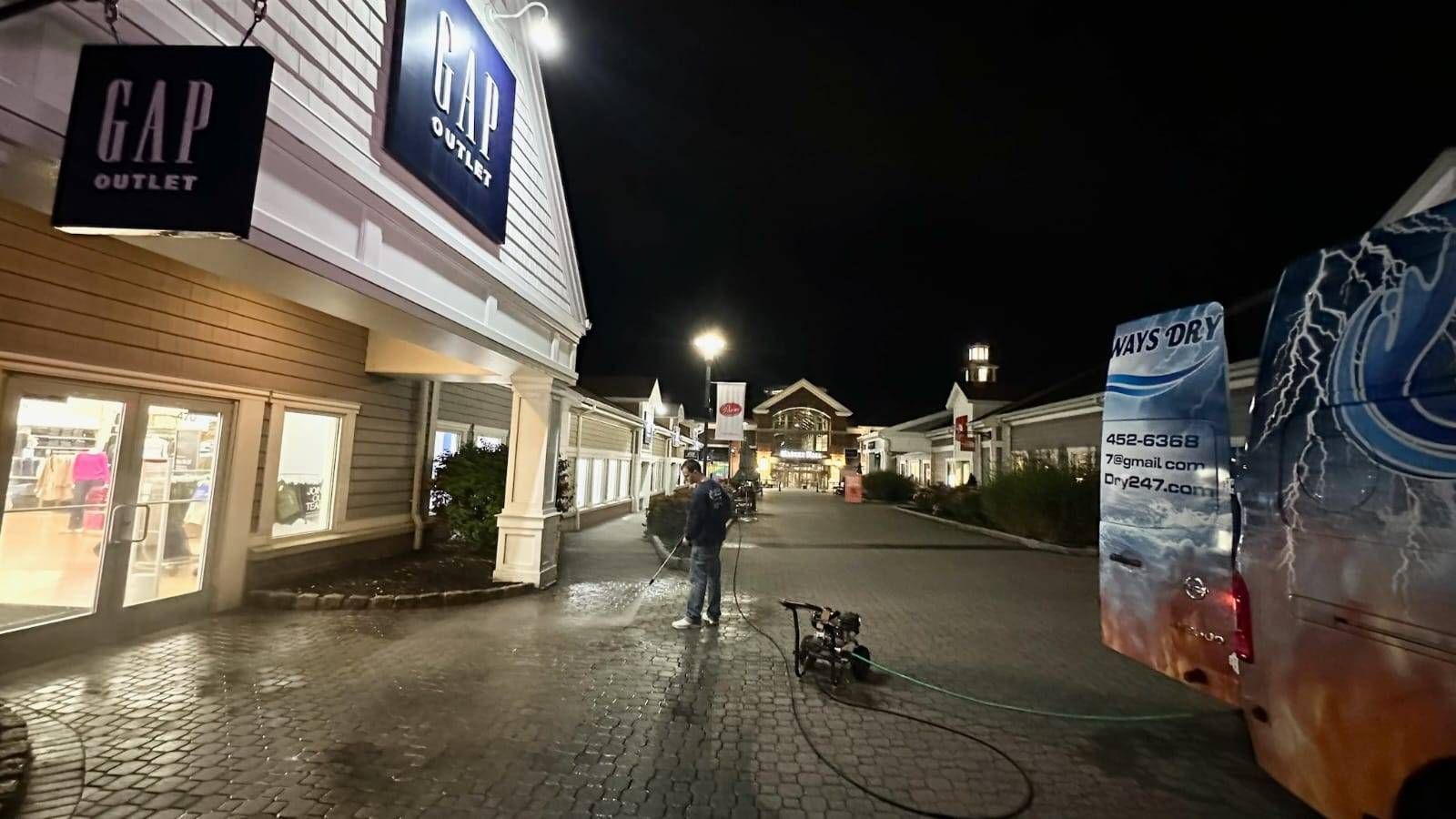 A person pressure washing the walkway in front of a Gap Outlet store at night in an outdoor shopping center.