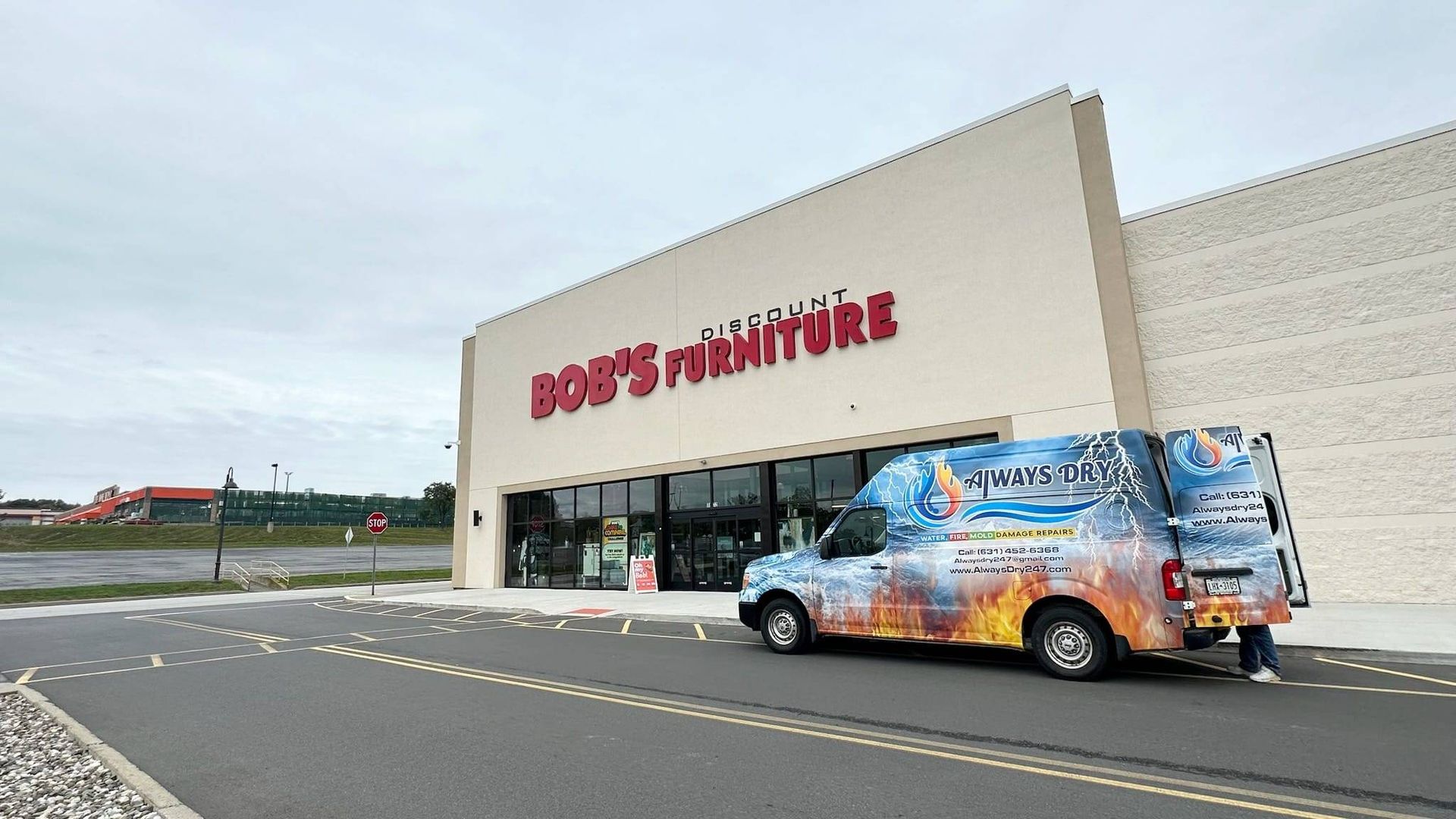 A van with a colorful, branded wrap parked in front of a Bob’s Discount Furniture store under an overcast sky.