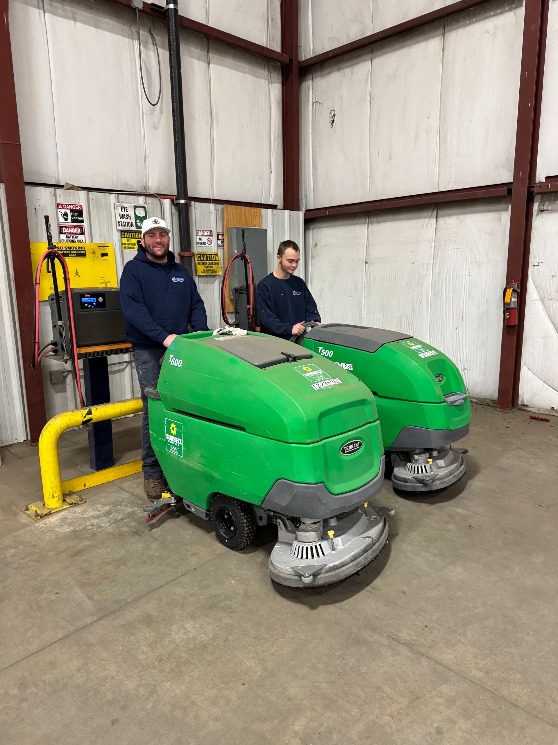 Two people stand in an industrial warehouse beside two large, bright green floor-scrubbing machines.
