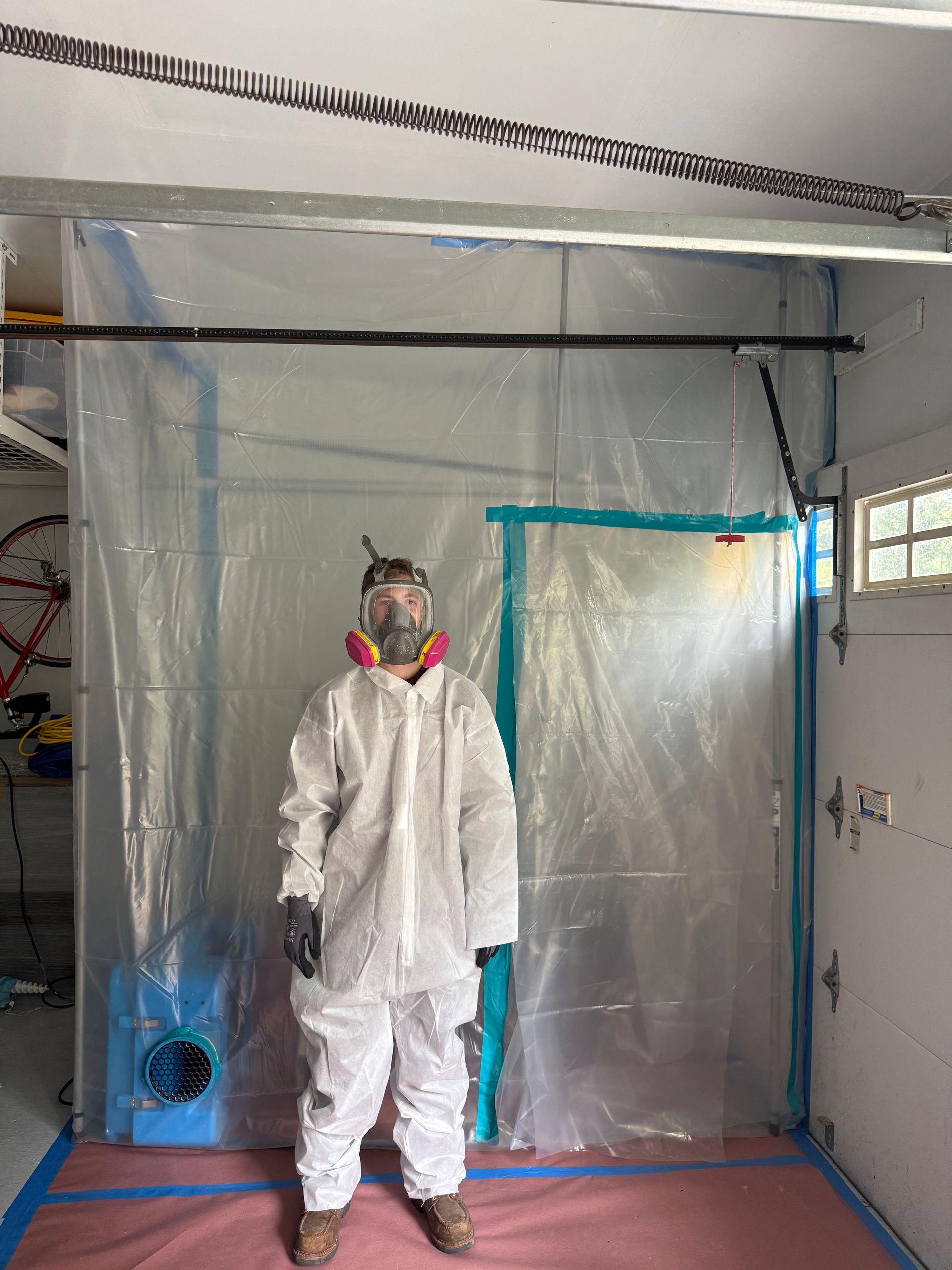 A person in a white hazmat suit and respirator stands in a garage inside a clear plastic containment enclosure.