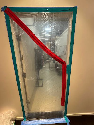 A doorway sealed with clear plastic sheeting and green painter's tape, featuring a diagonal red adhesive zipper.