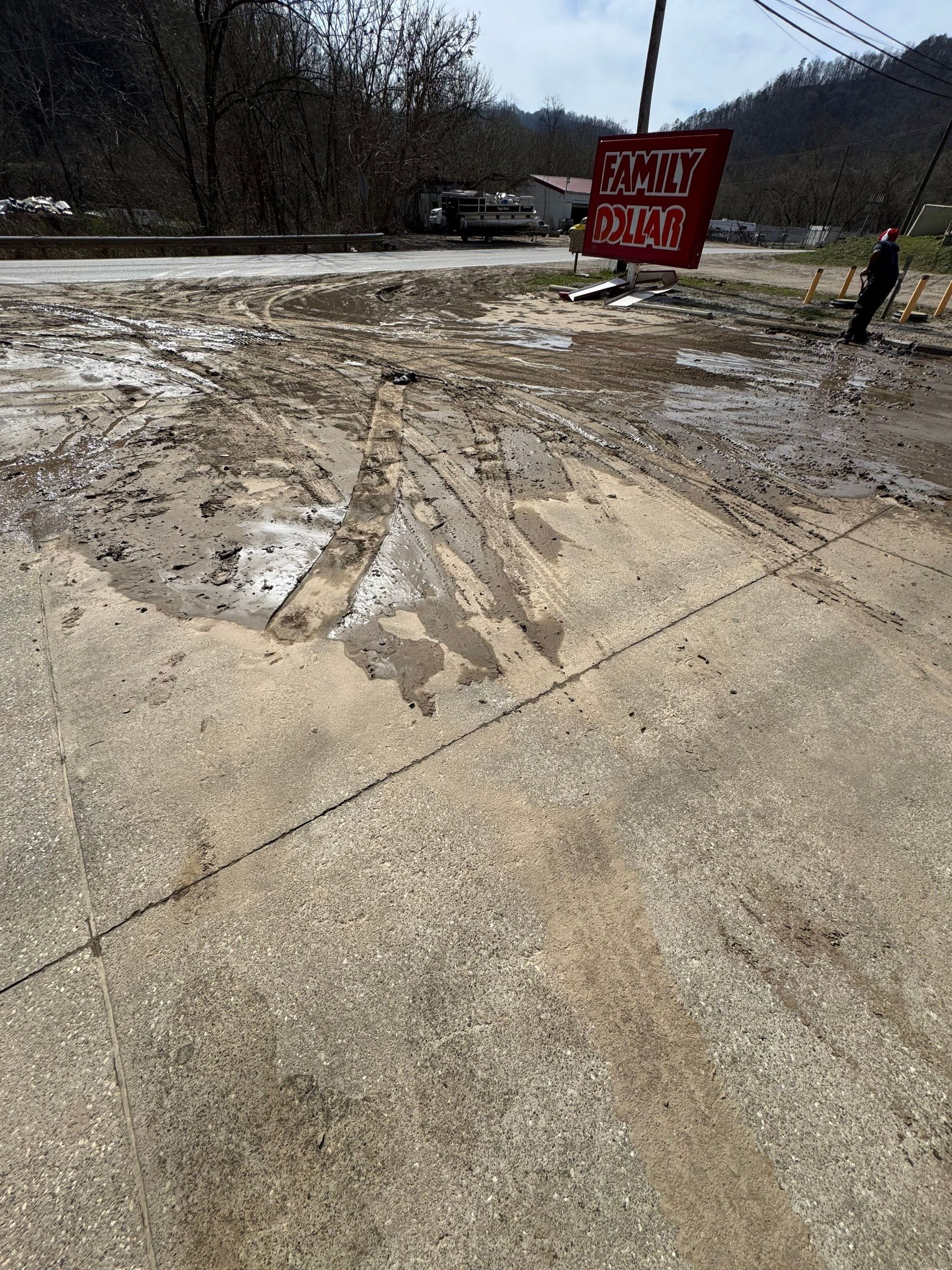 A red sign stands in a muddy, debris-covered parking lot on an overcast day with hills in the background.