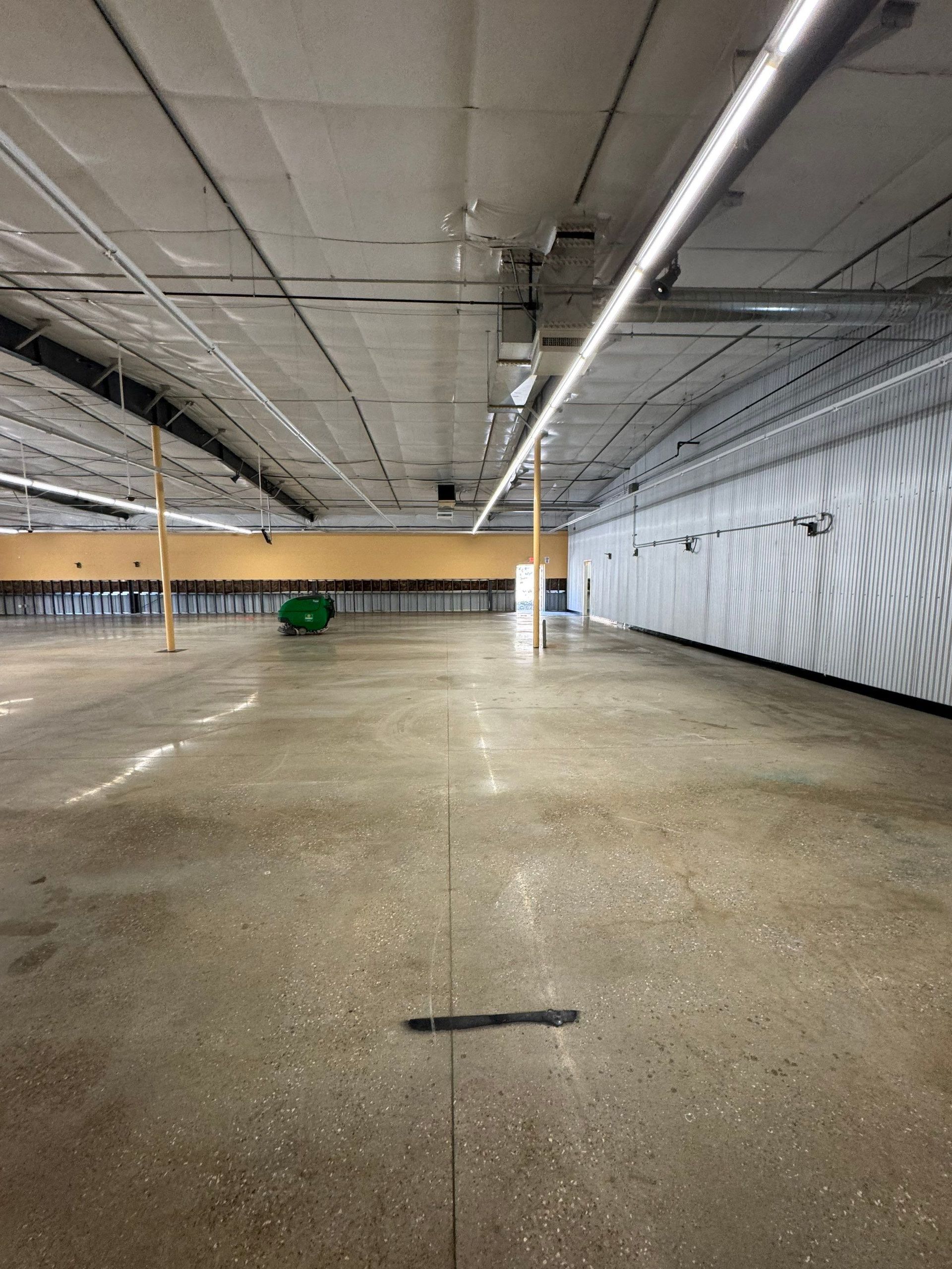 A wide, empty retail space with polished concrete floors, a drop ceiling, exposed lighting, and a single green bin.
