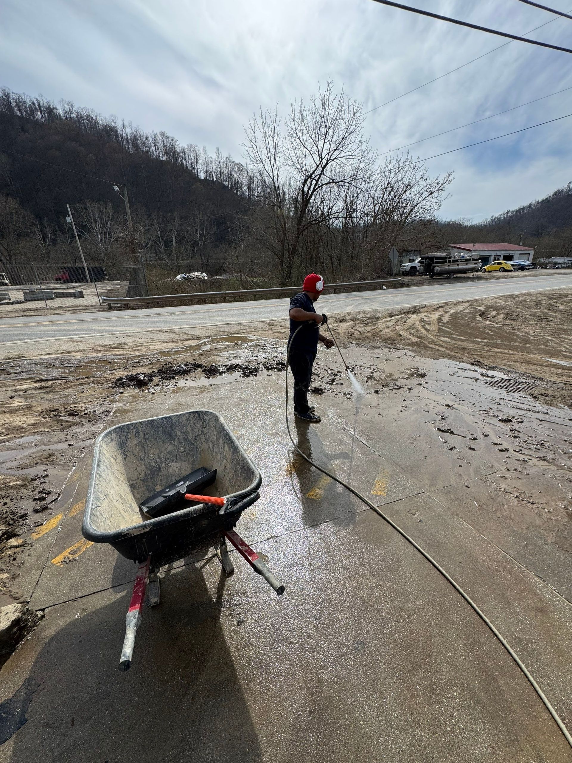 A person in a red cap stands on a mud-covered surface next to a wheelbarrow, with a debris-strewn landscape