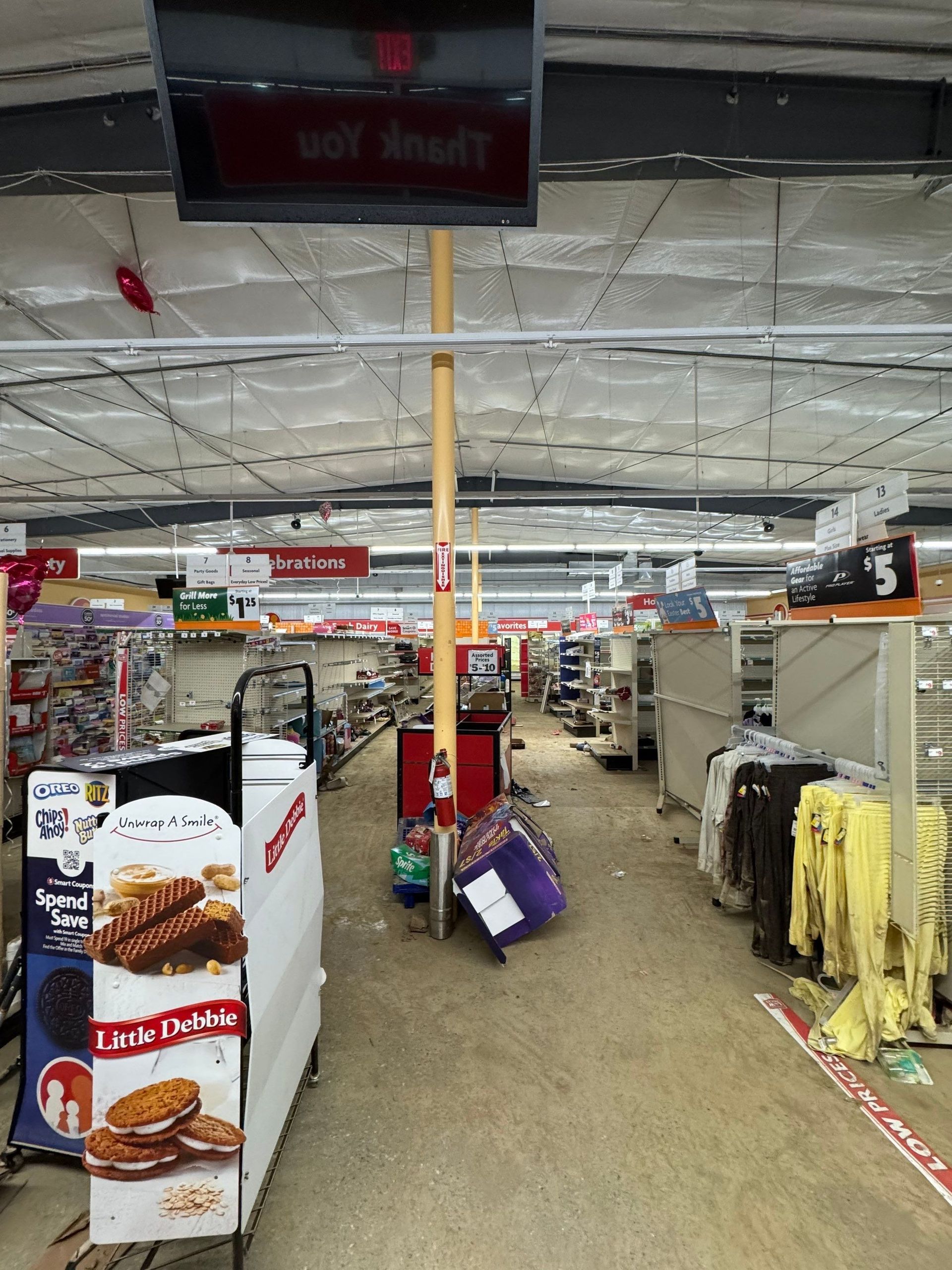 A dimly lit, cluttered store aisle featuring shelving units, a display of cookie packages, and racks of hanging clothing.