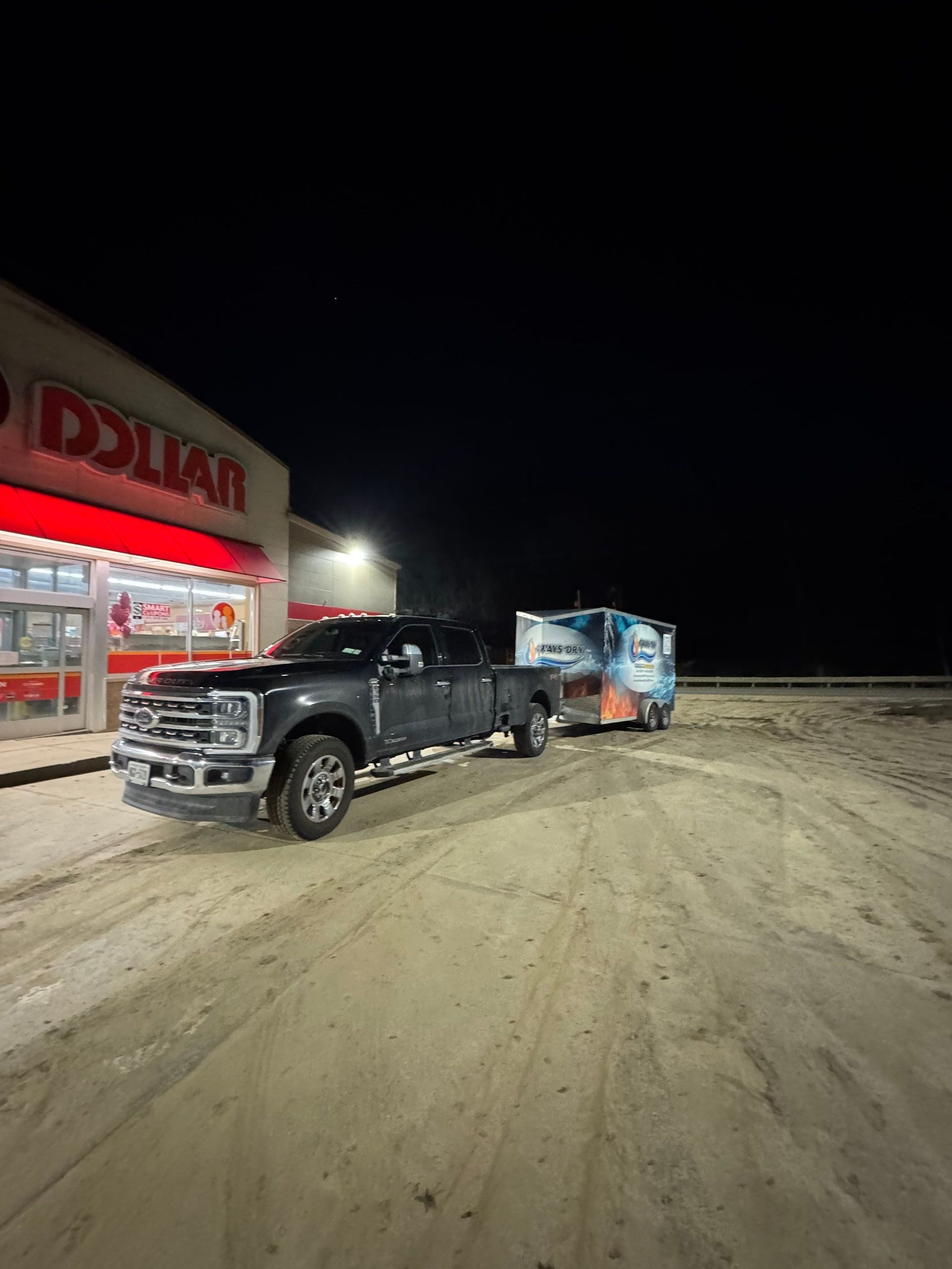 A dark pickup truck towing a trailer parked in the dirt lot in front of a Dollar General store at night.