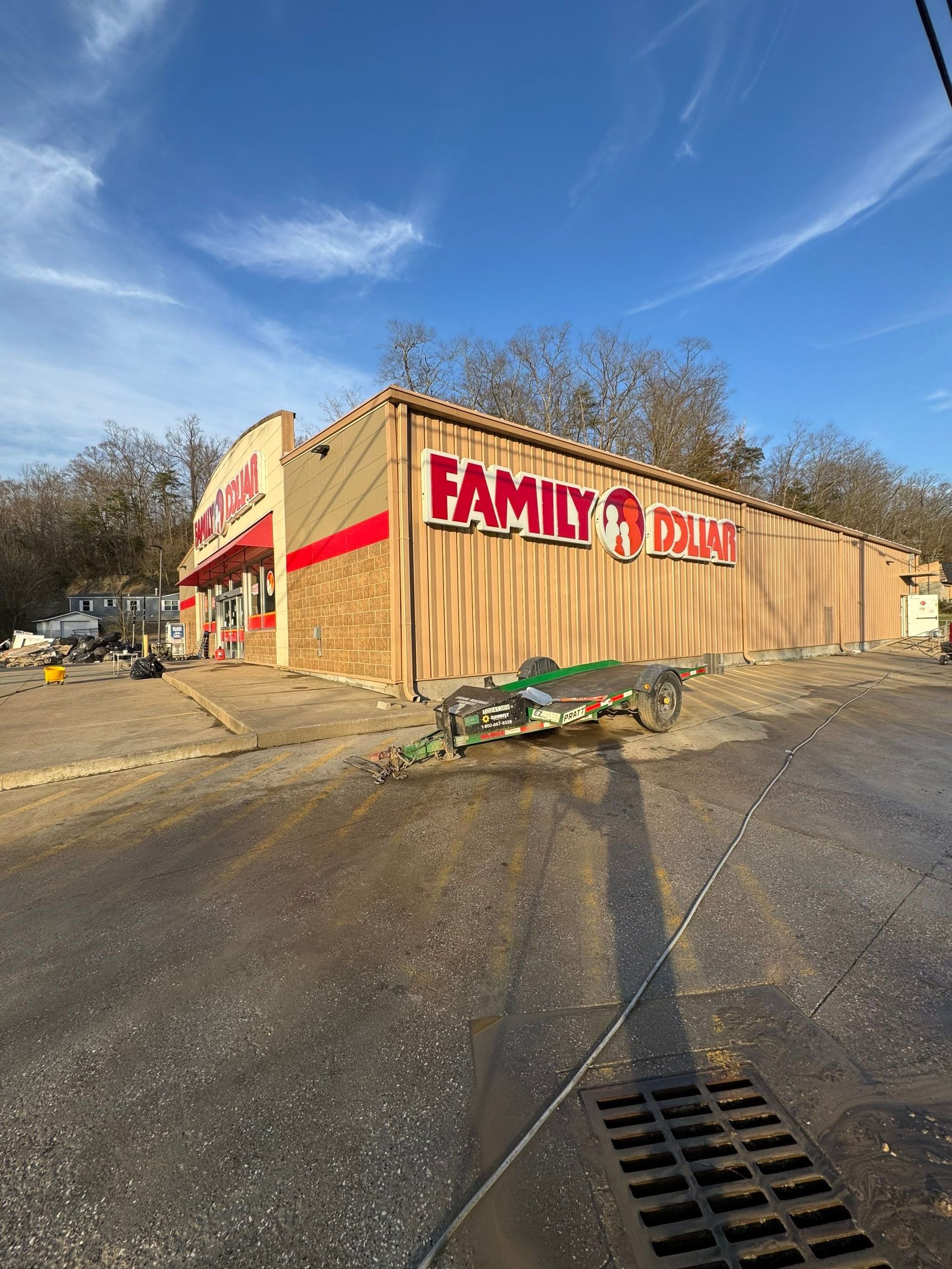A Family Dollar store building with boarded-up windows and a small trailer parked in the paved lot under a blue sky.