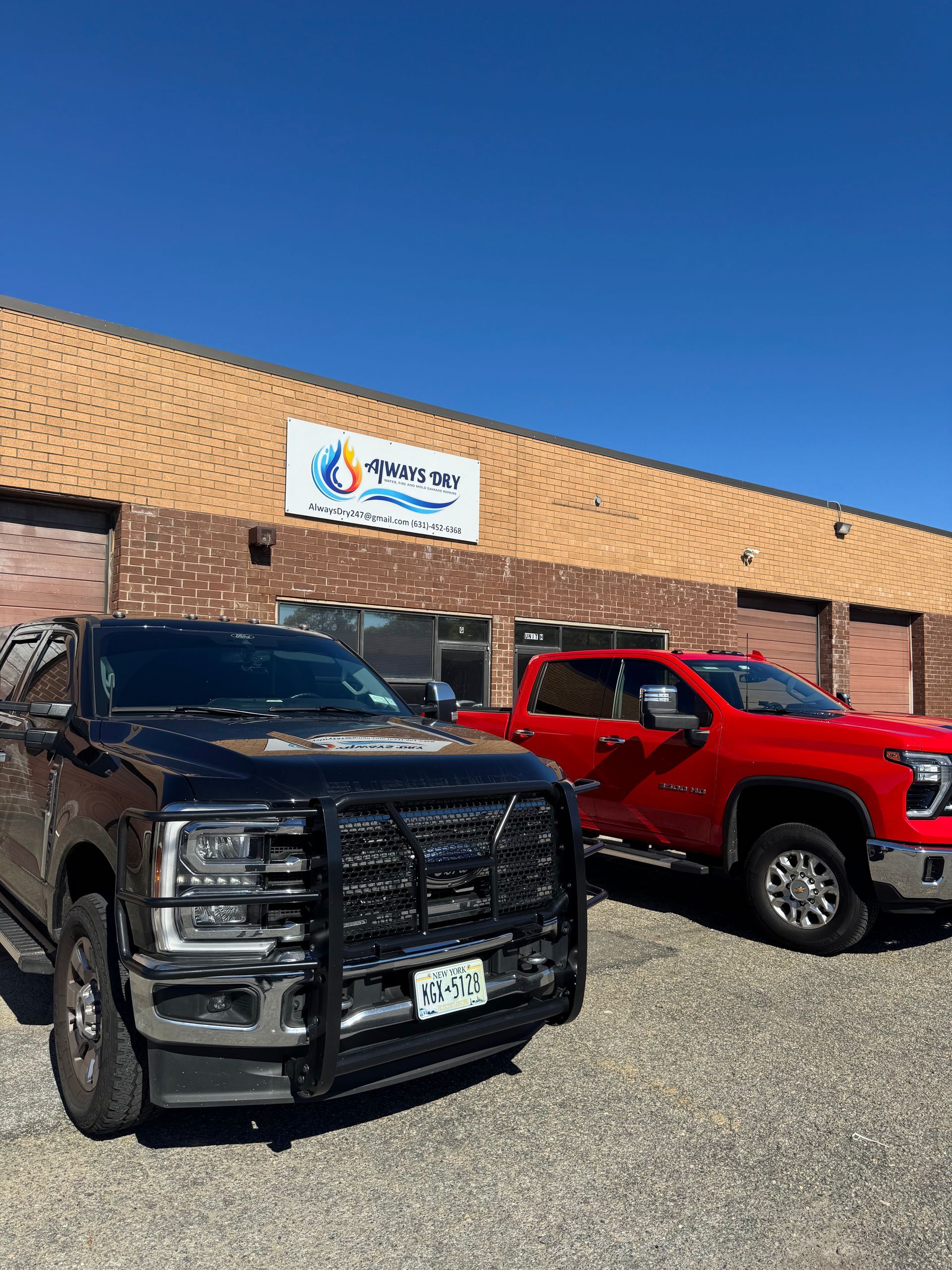 A black truck and a red truck parked in front of a tan building with a blue and white business sign.