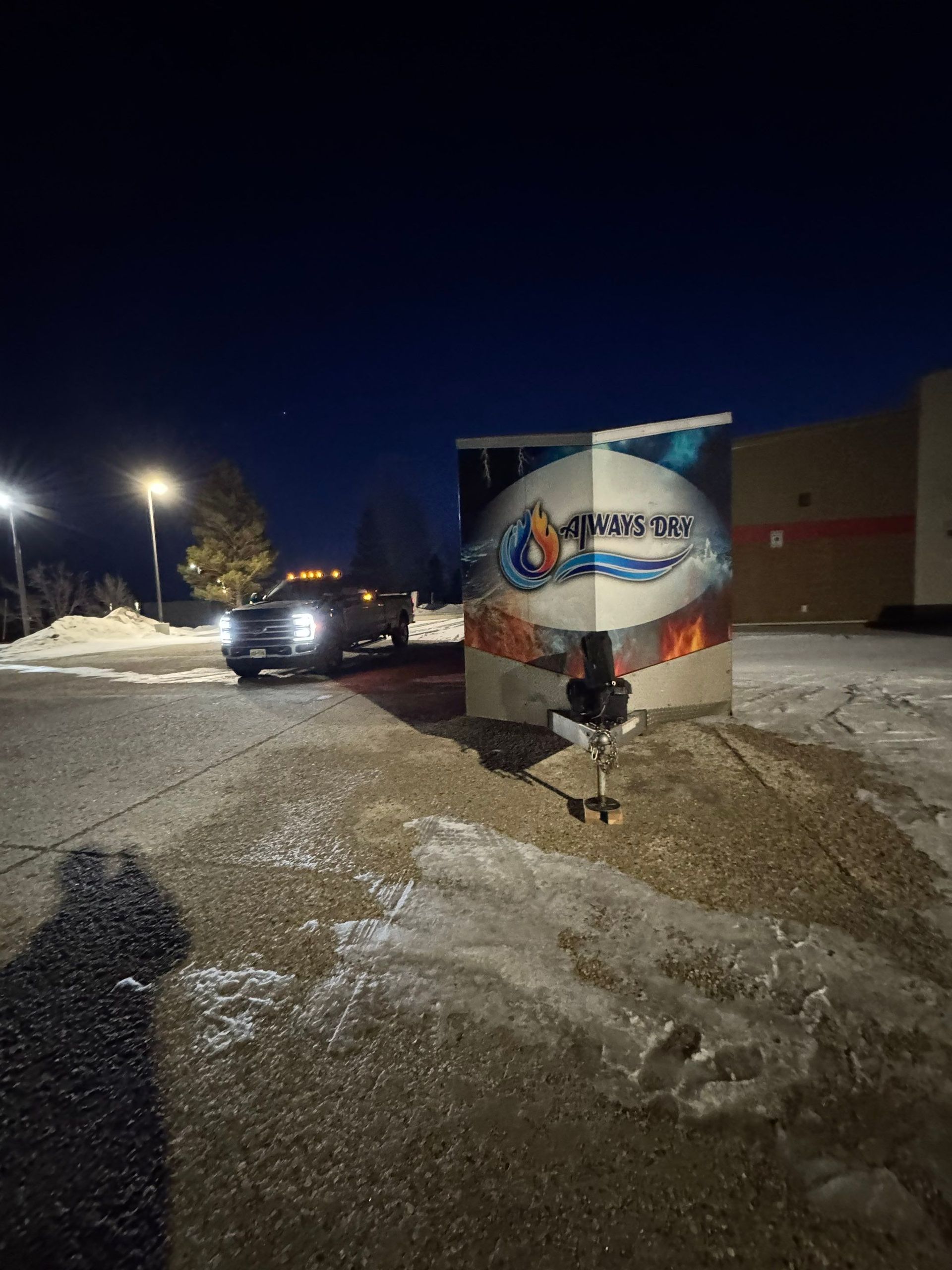 A person sits on a stool next to a mural depicting a flaming gas logo in a dark, snowy parking lot at night.