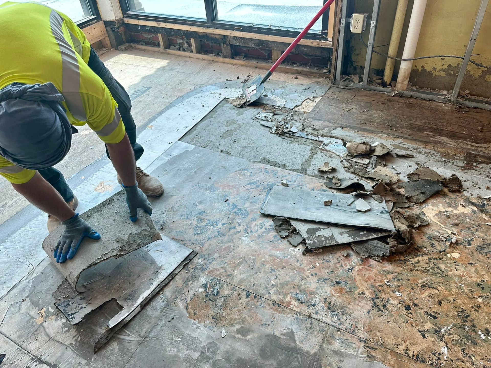 A construction worker in a high-visibility yellow shirt scrapes old tile flooring from an indoor subfloor.