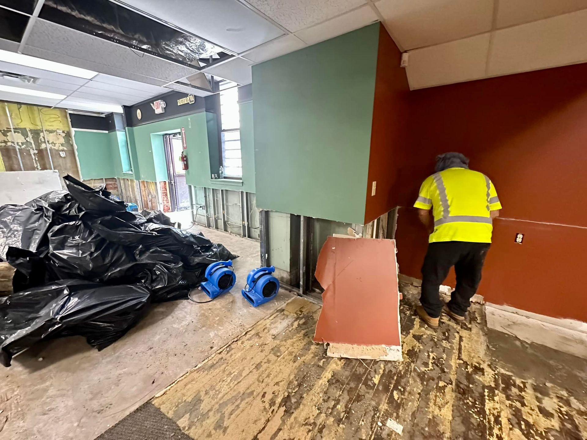 A worker in a high-visibility vest stands in a room undergoing renovation, featuring black debris bags and blue fans.