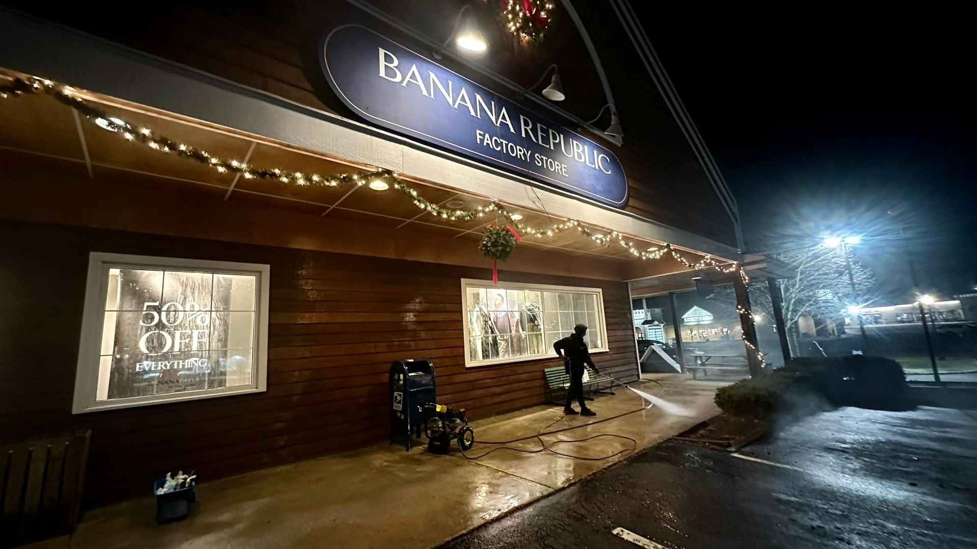 A Banana Republic Factory Store at night, featuring festive holiday lighting and a person sweeping the entrance walkway.