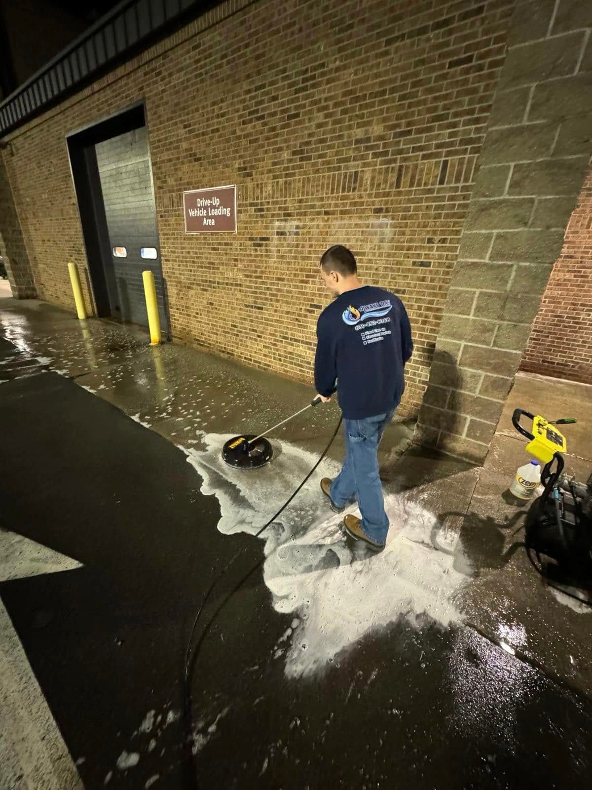 A person in a dark uniform uses a surface cleaner attached to a pressure washer on a concrete walkway by a brick wall.
