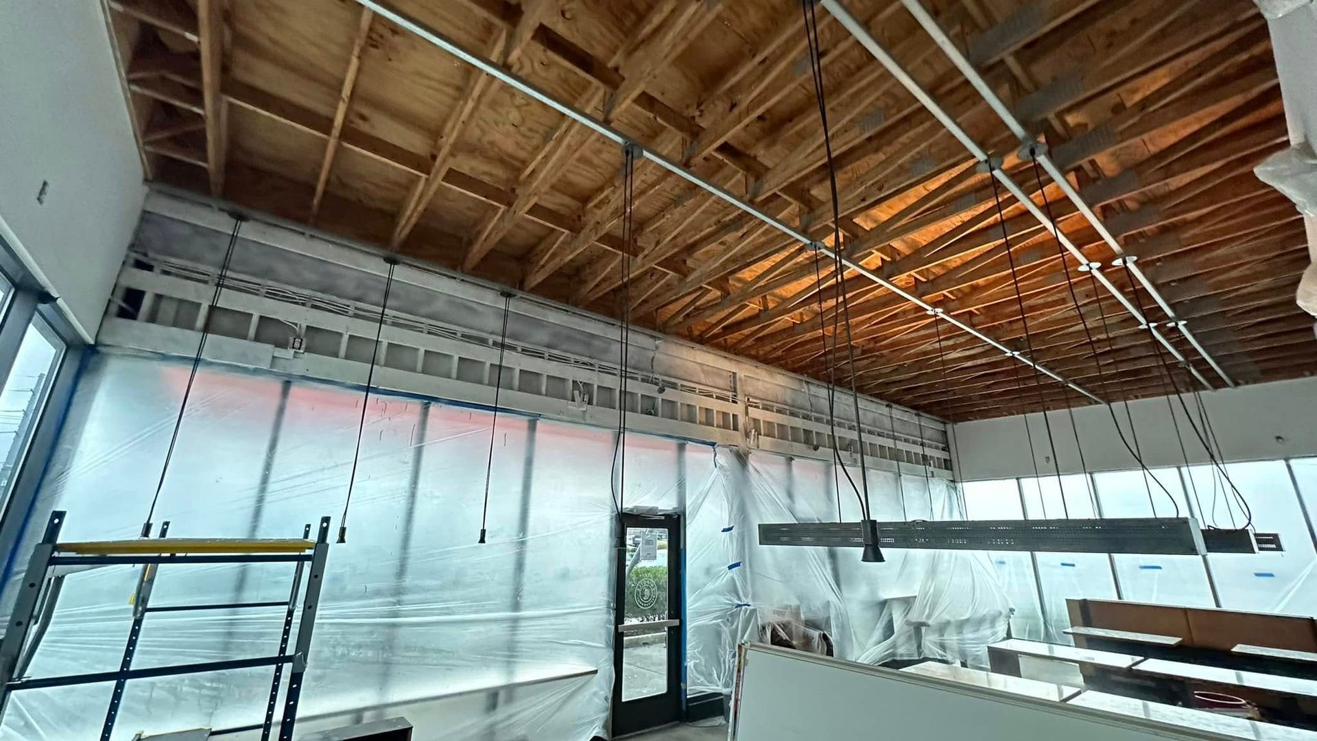 An interior construction site featuring exposed wood ceiling joists, white plastic-covered walls, and hanging light fixtures.