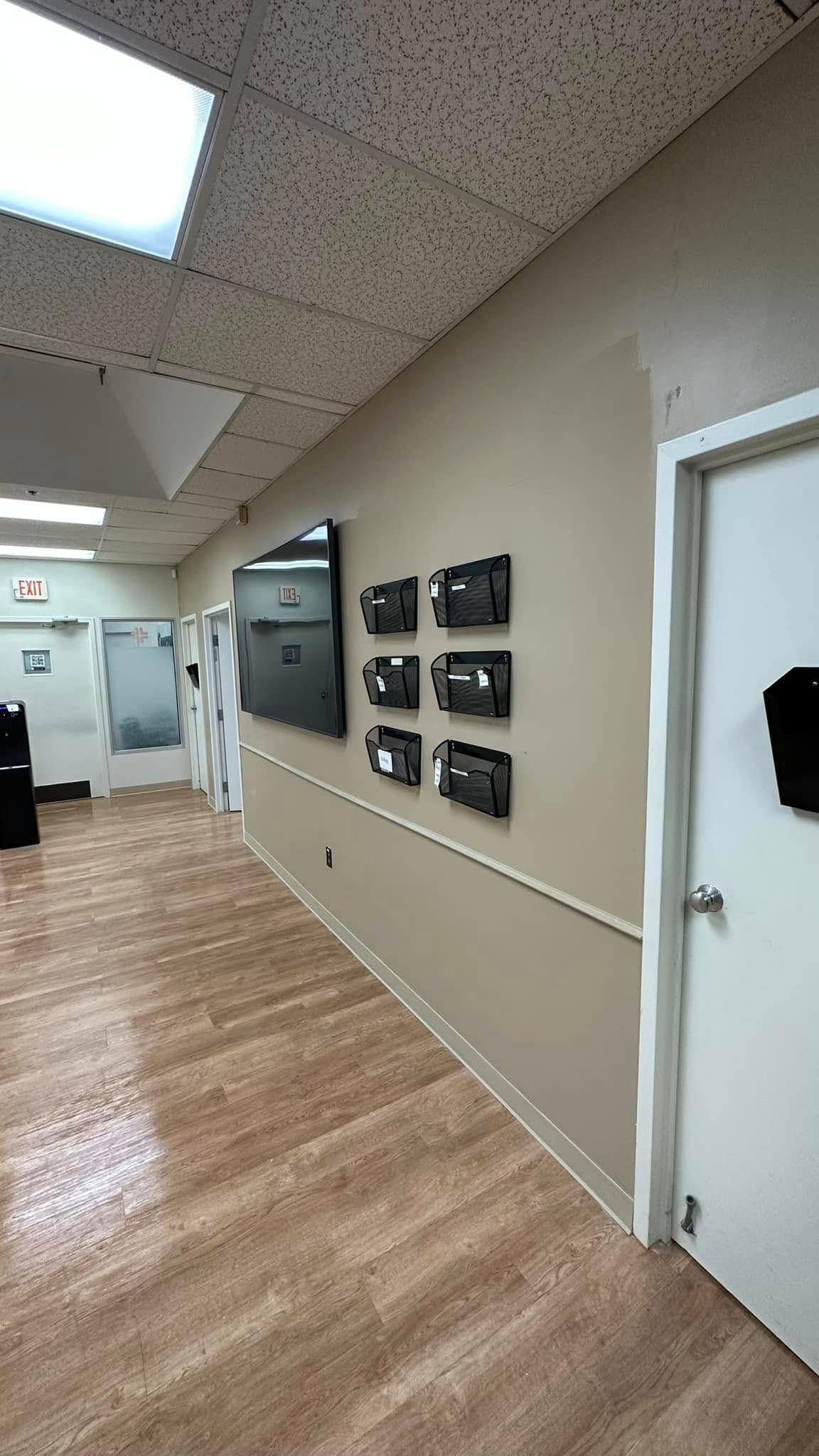 A hallway with wood-look flooring, a beige wall with a row of six small black boxes, a flat-screen TV, and a white door.