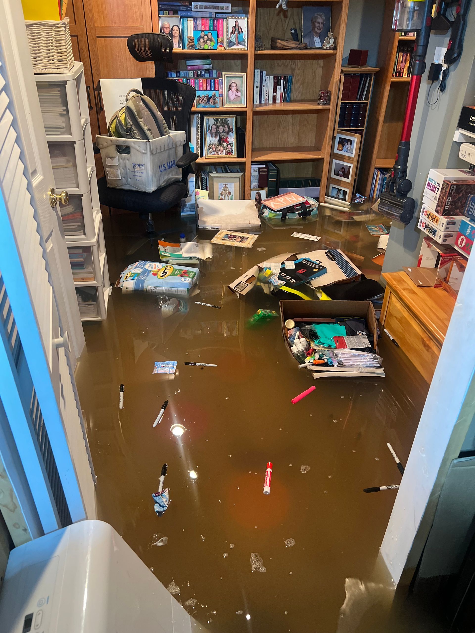 A flooded room with brown water covering the floor, submerging various items including pens, boxes, and a chair.