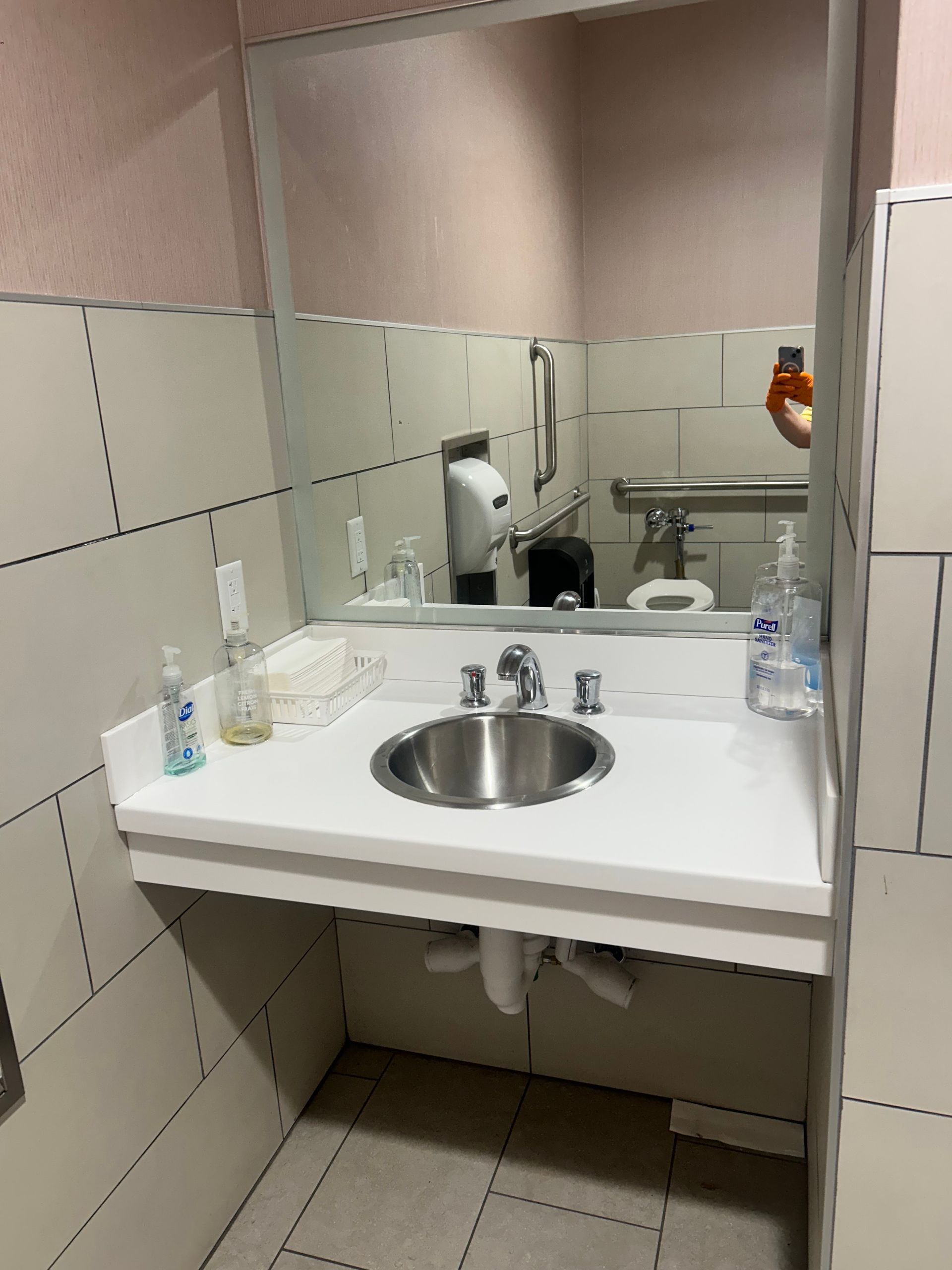 A wall-mounted bathroom sink with a metal basin, mirror, and soap dispensers, showing a person taking the photo.