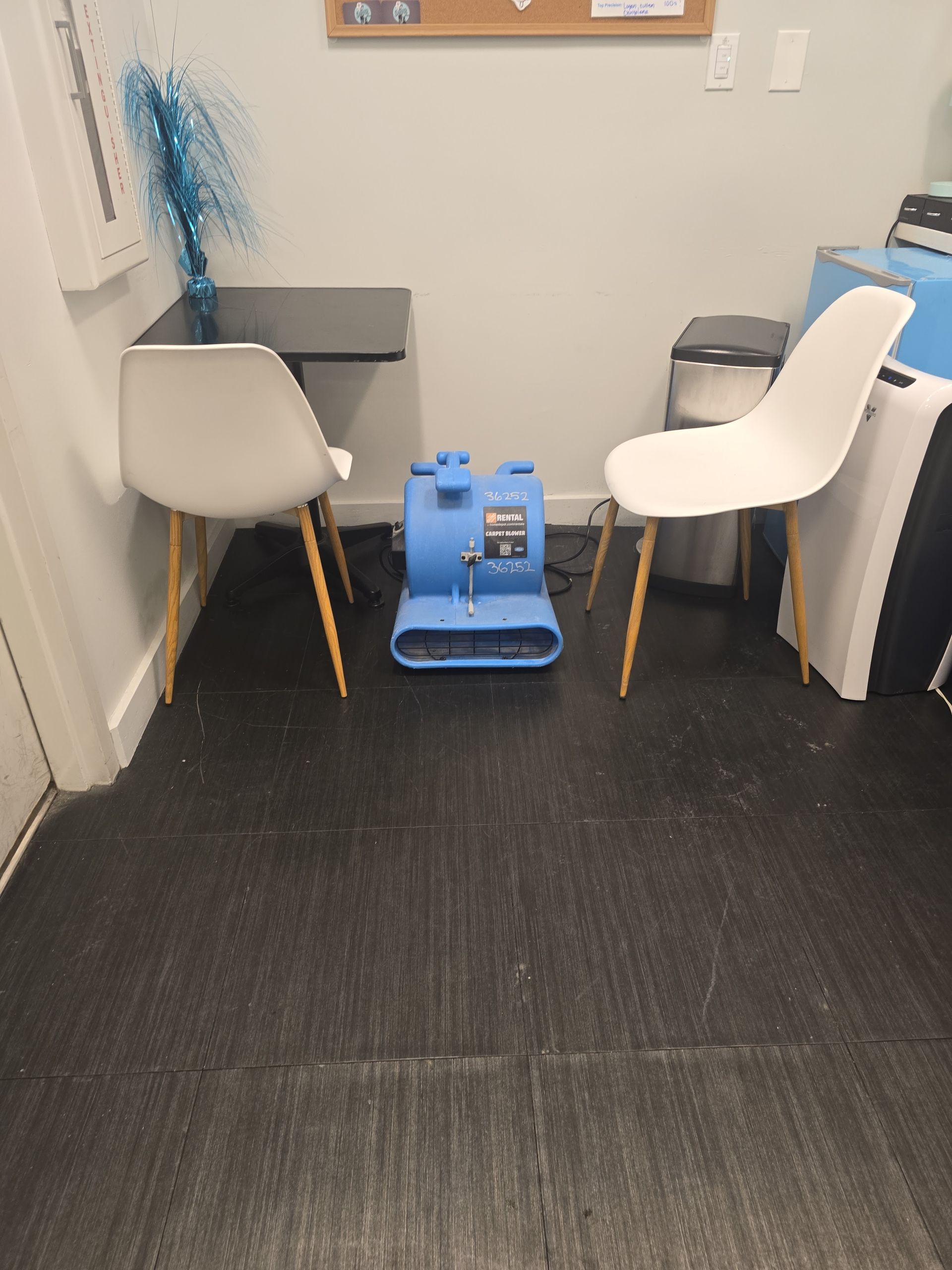 A blue air mover sits on a dark floor between two white chairs at a small black table in an office break area.