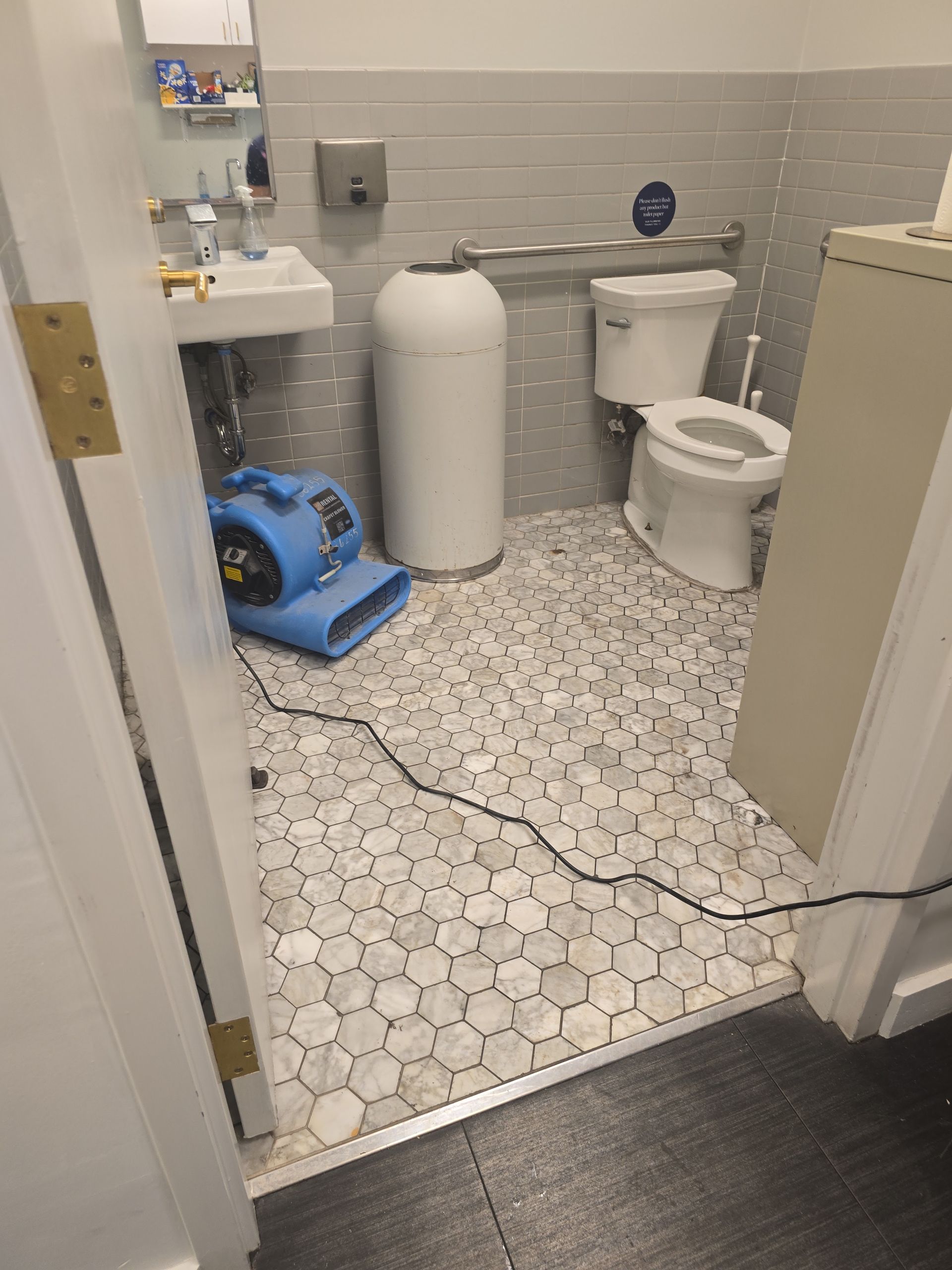 A bathroom with a sink, toilet, and blue industrial floor dryer running near the sink, indicating recent water damage.