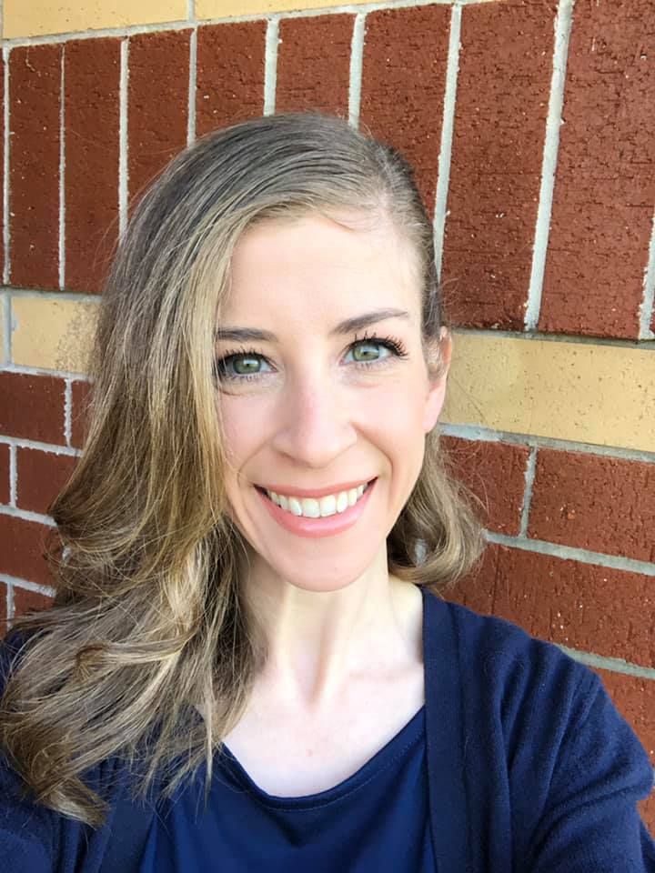 Woman with long wavy hair smiles in front of a brick wall, wearing a dark blue top and cardigan.