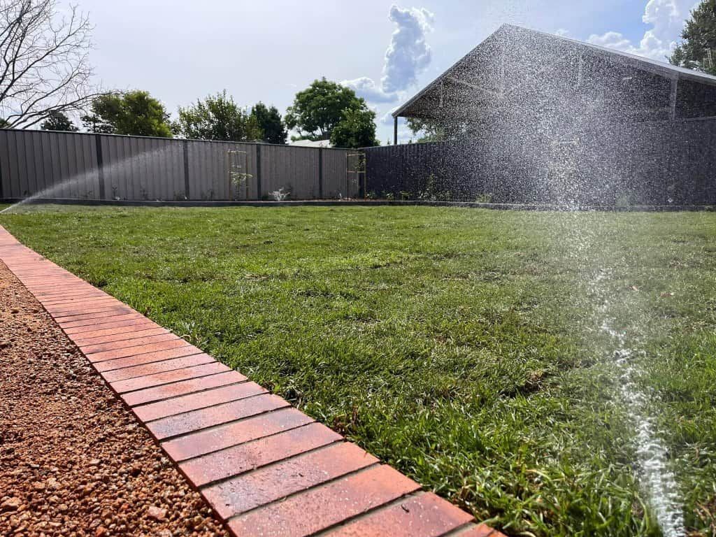 Sprinkler watering a green lawn in a backyard with a brick border, fence, and house in the background — Darlington Landscaping in Dubbo, NSW