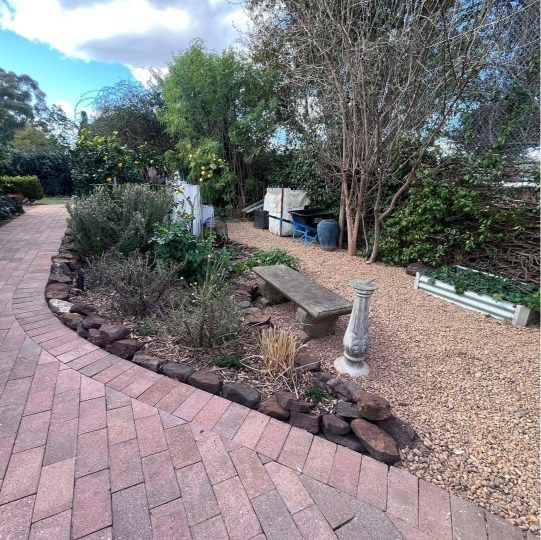 Brick path leads to a garden with stone bench, plants, and gravel pathway. Trees and blue sky visible — Darlington Landscaping in Dubbo, NSW