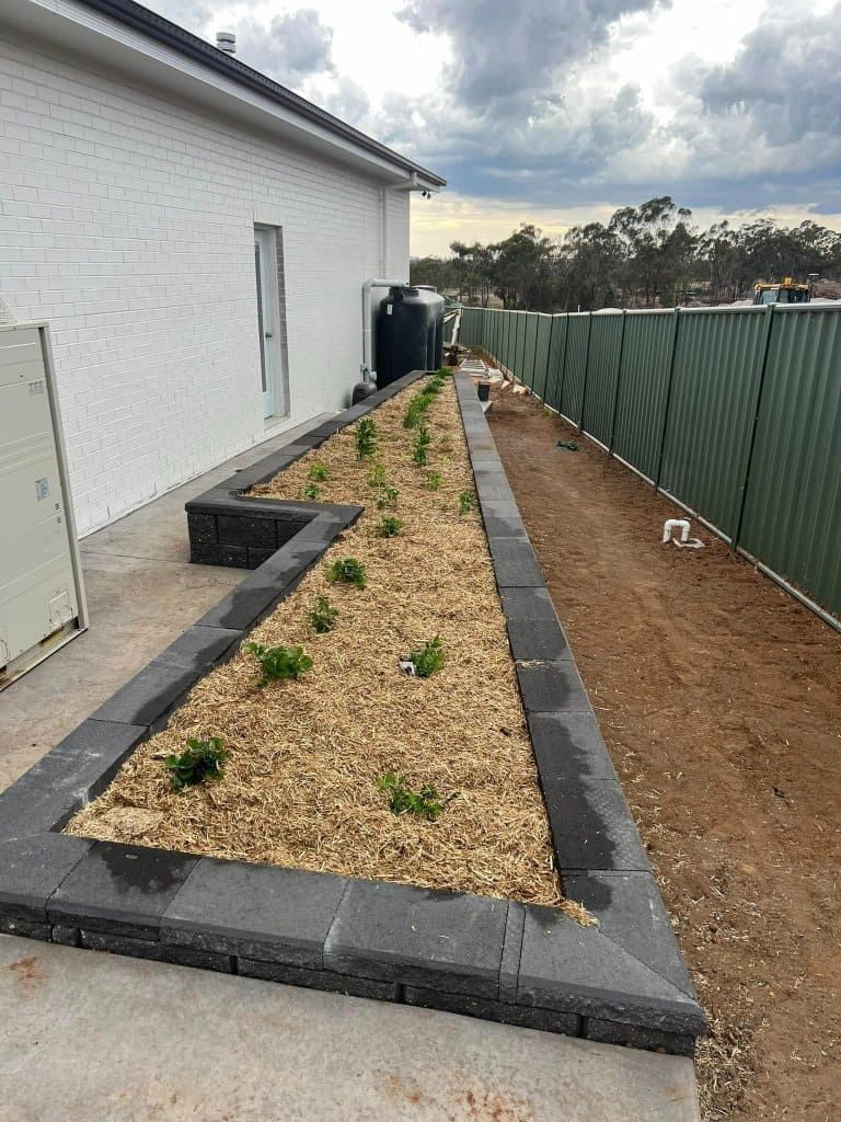 A long, narrow garden bed along a white building with a green fence. The bed has mulch, young plants, and dark stone edging — Darlington Landscaping in Dubbo, NSW
