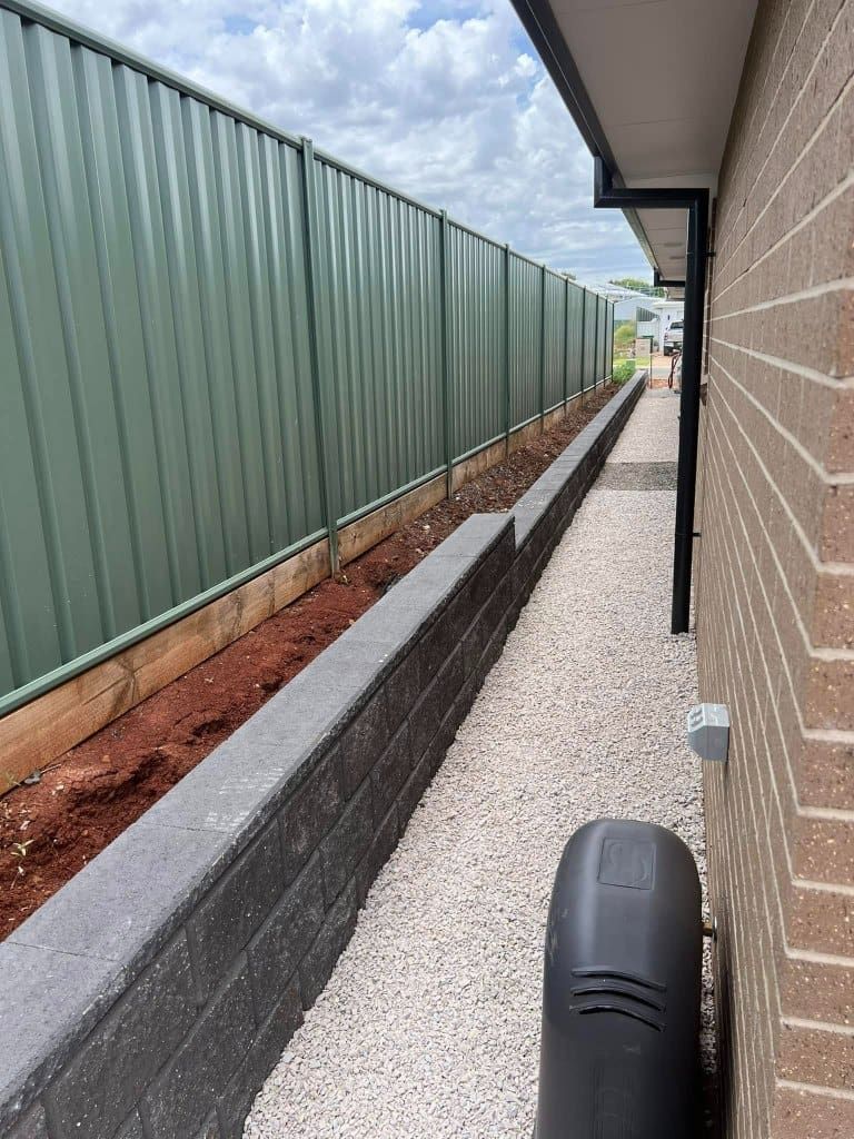 A narrow side yard with a grey retaining wall, gravel path, green fence, and brick house — Darlington Landscaping in Dubbo, NSW