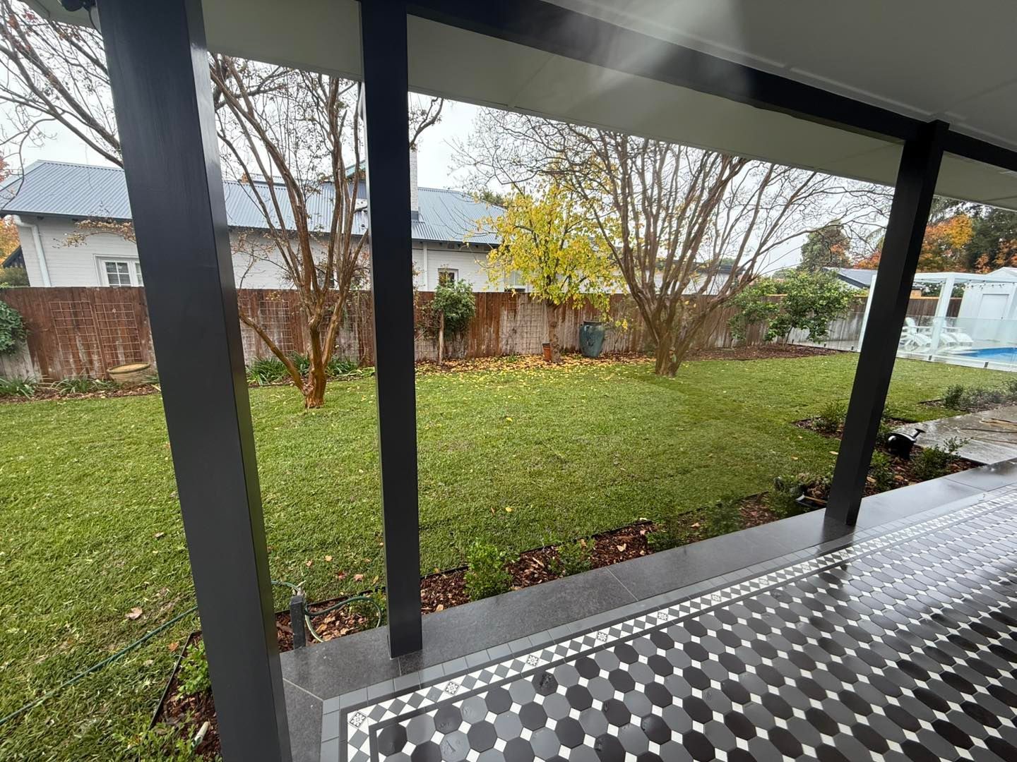 View from a covered patio to a green backyard with trees, houses, and a patterned floor — Darlington Landscaping in Dubbo, NSW