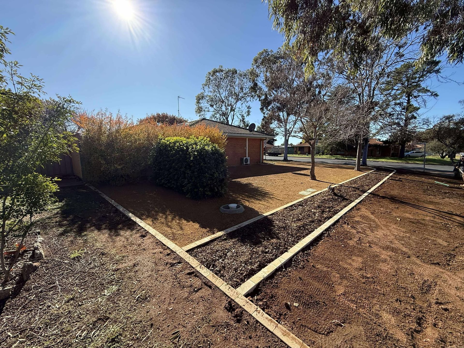 A sunny backyard with freshly laid borders. Brown soil, green and brown bushes, and a distant house — Darlington Landscaping in Nyngan, NSW