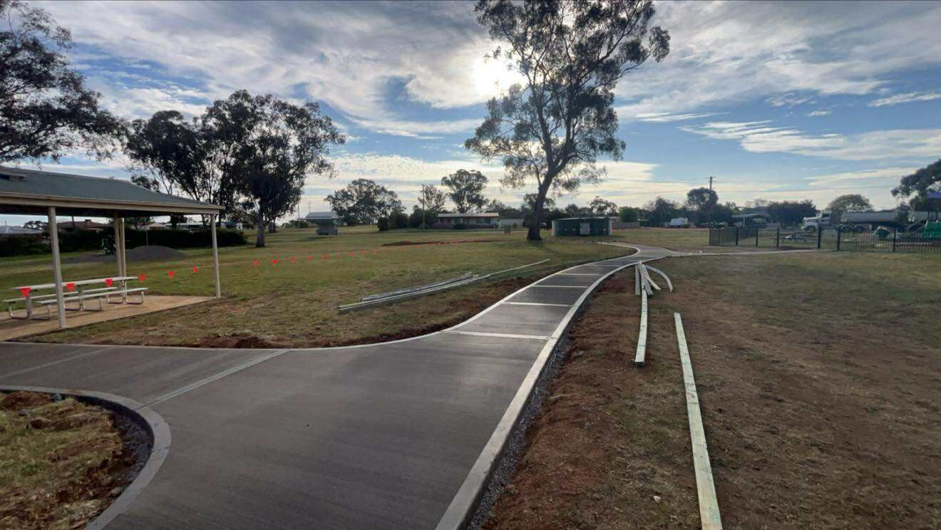 A paved path curves through a park, passing a shelter and grassy area, under a cloudy sky — Darlington Landscaping in Parkes, NSW