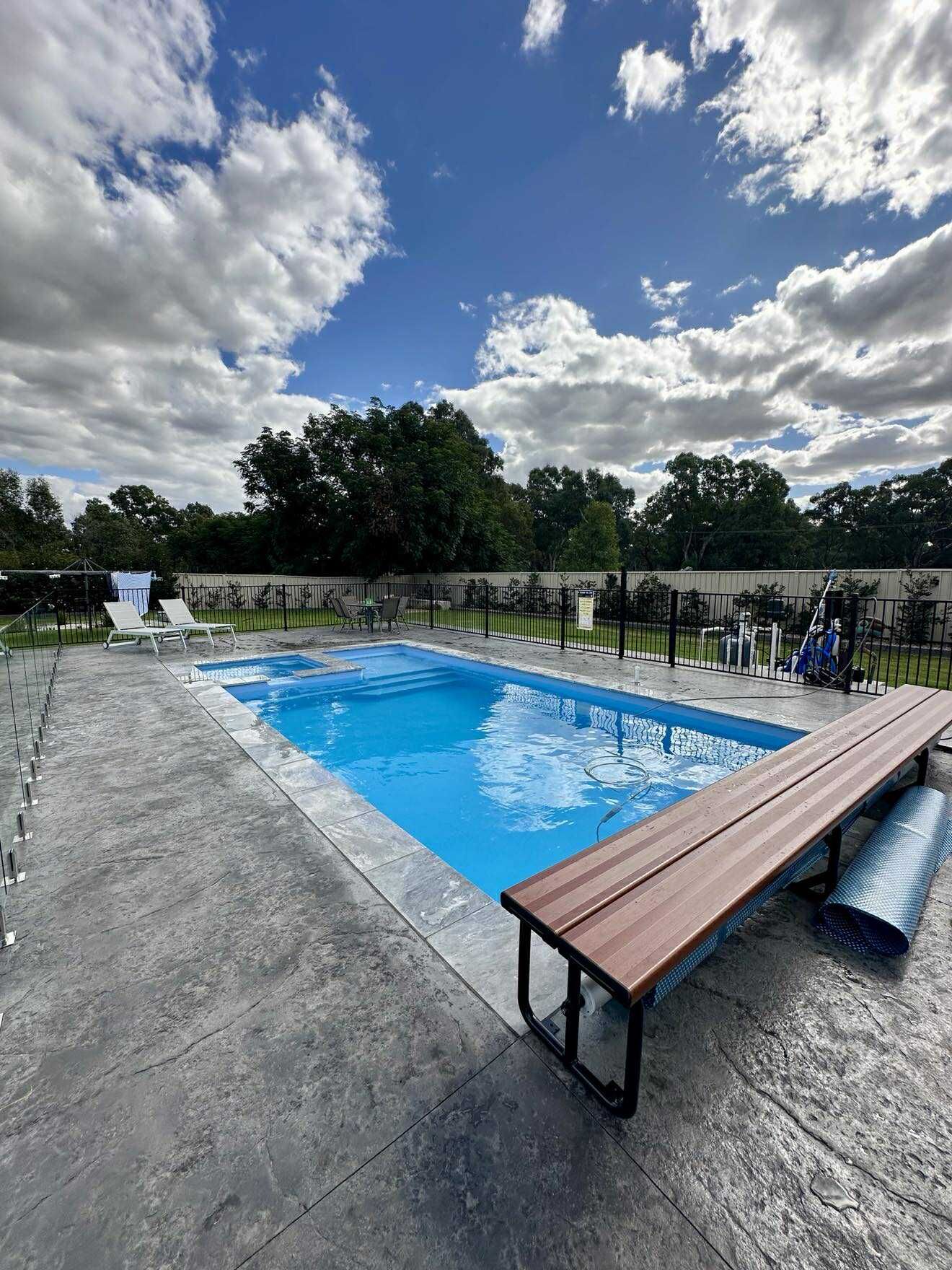 A rectangular pool with blue water, surrounded by grey concrete. A brown bench and a cloudy sky are also visible — Darlington Landscaping in Dubbo, NSW