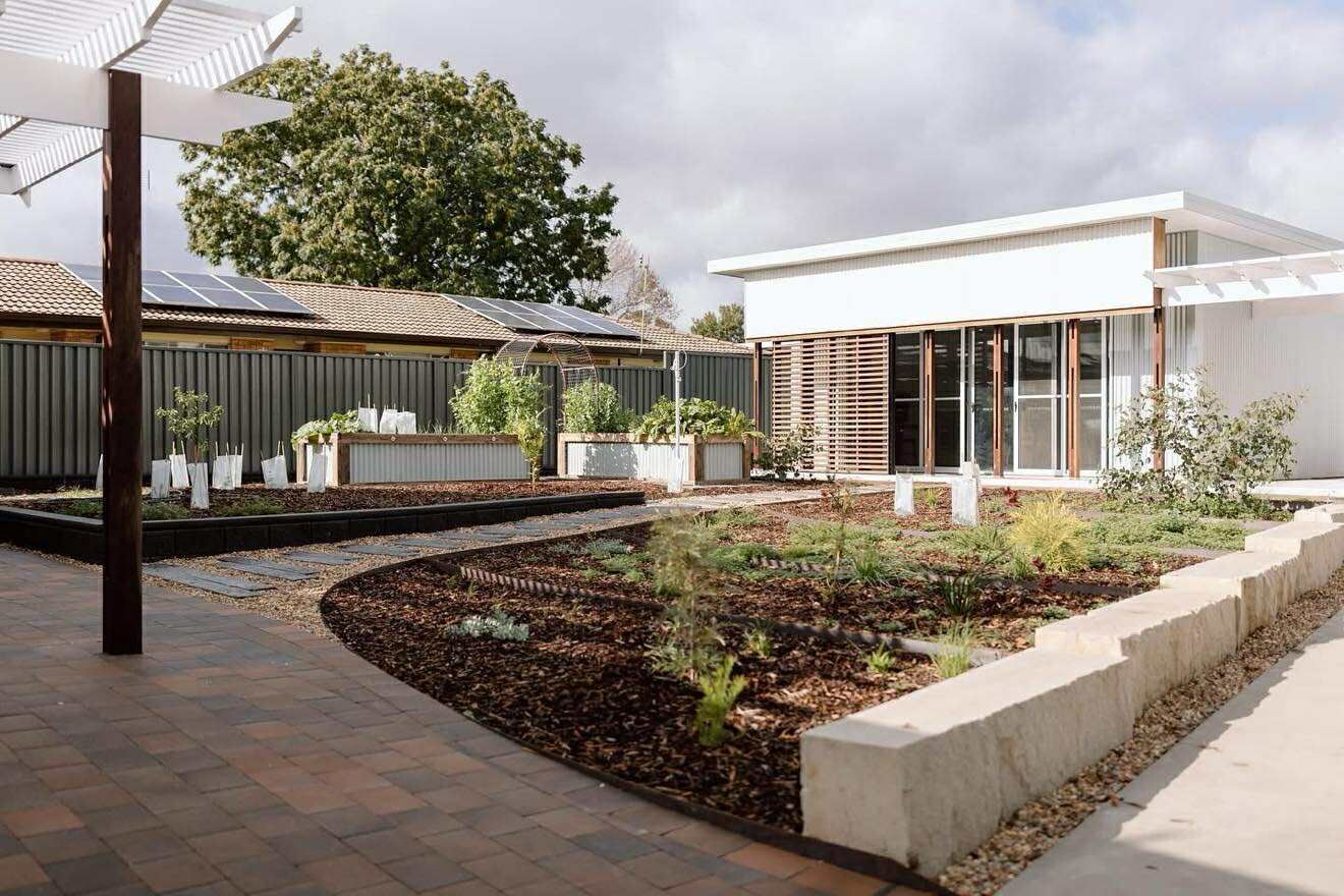 Garden with flowerbeds, gravel paths, and white building. Pergola and solar panels visible. Sunny day — Darlington Landscaping in Dubbo, NSW