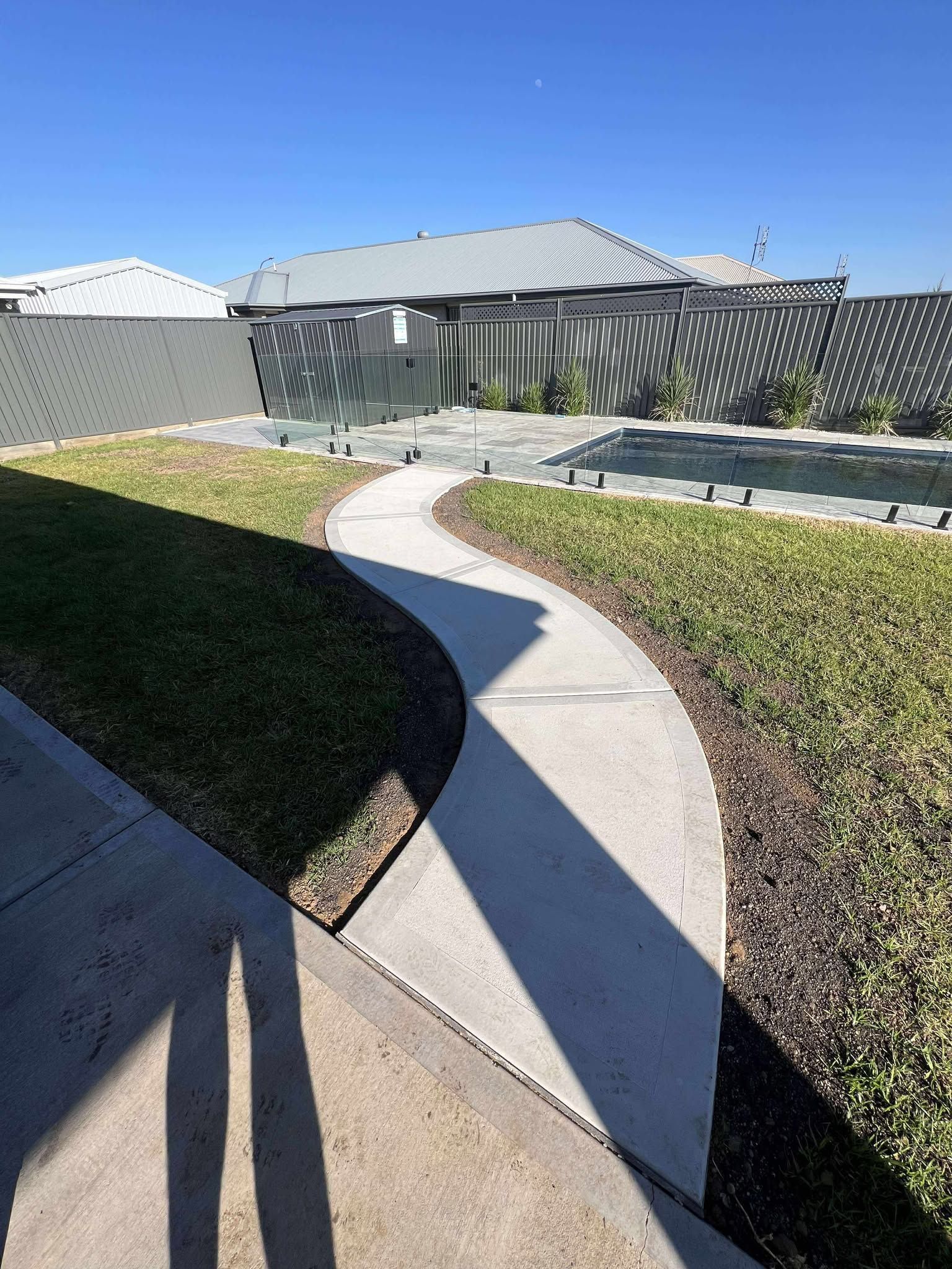 Concrete path curves through grassy yard, leading to a pool with glass fencing. Sunny day — Darlington Landscaping in Dubbo, NSW