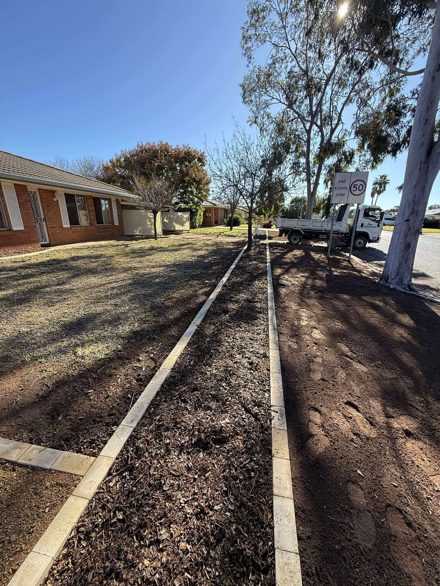 Pathway with wood chips, bordered by concrete, next to a building and a truck on a sunny day — Darlington Landscaping in Mudgee, NSW