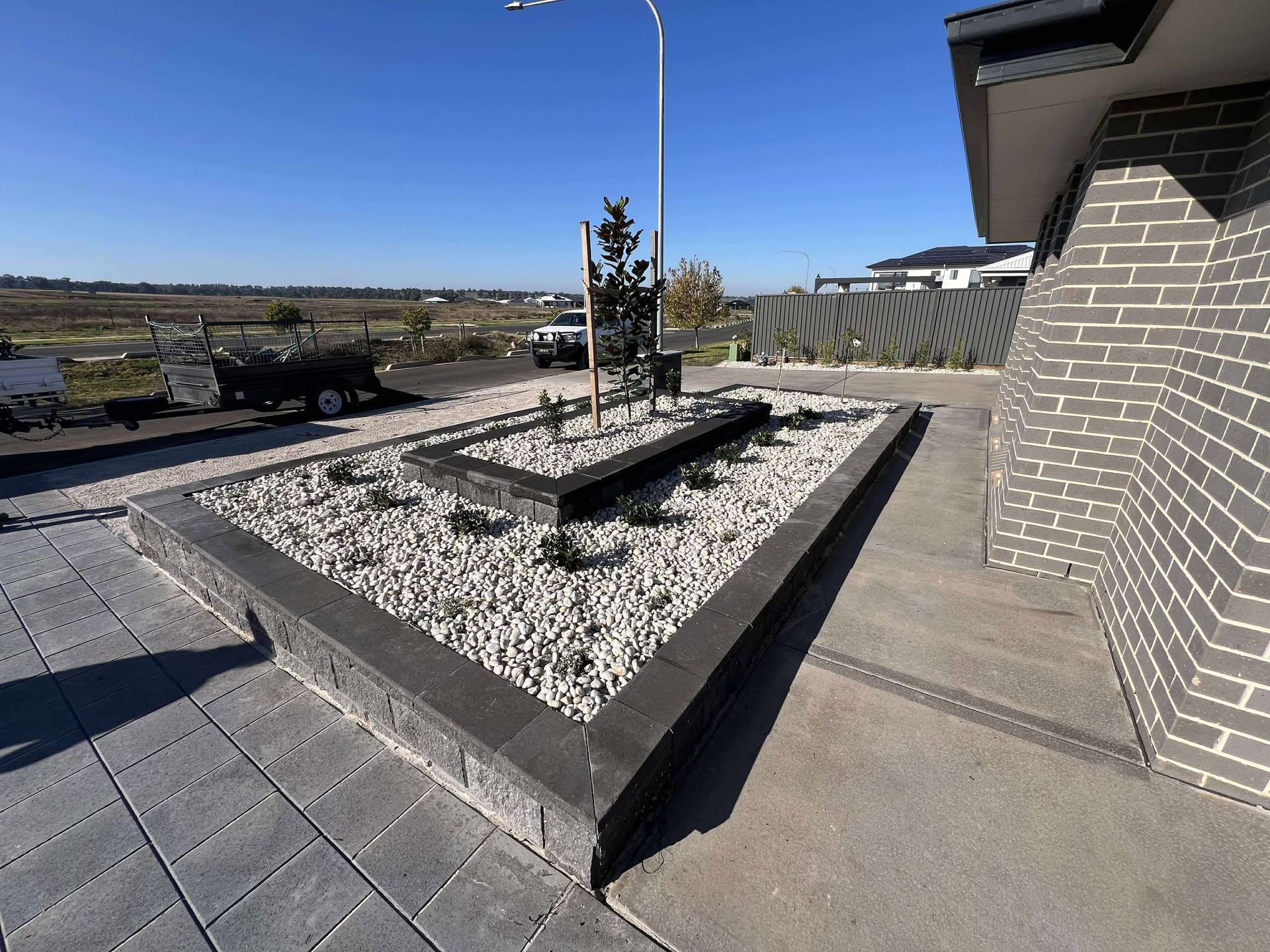 Triangular garden bed with grey bricks and white pebbles next to a grey brick house on a sunny day — Darlington Landscaping in Dubbo, NSW