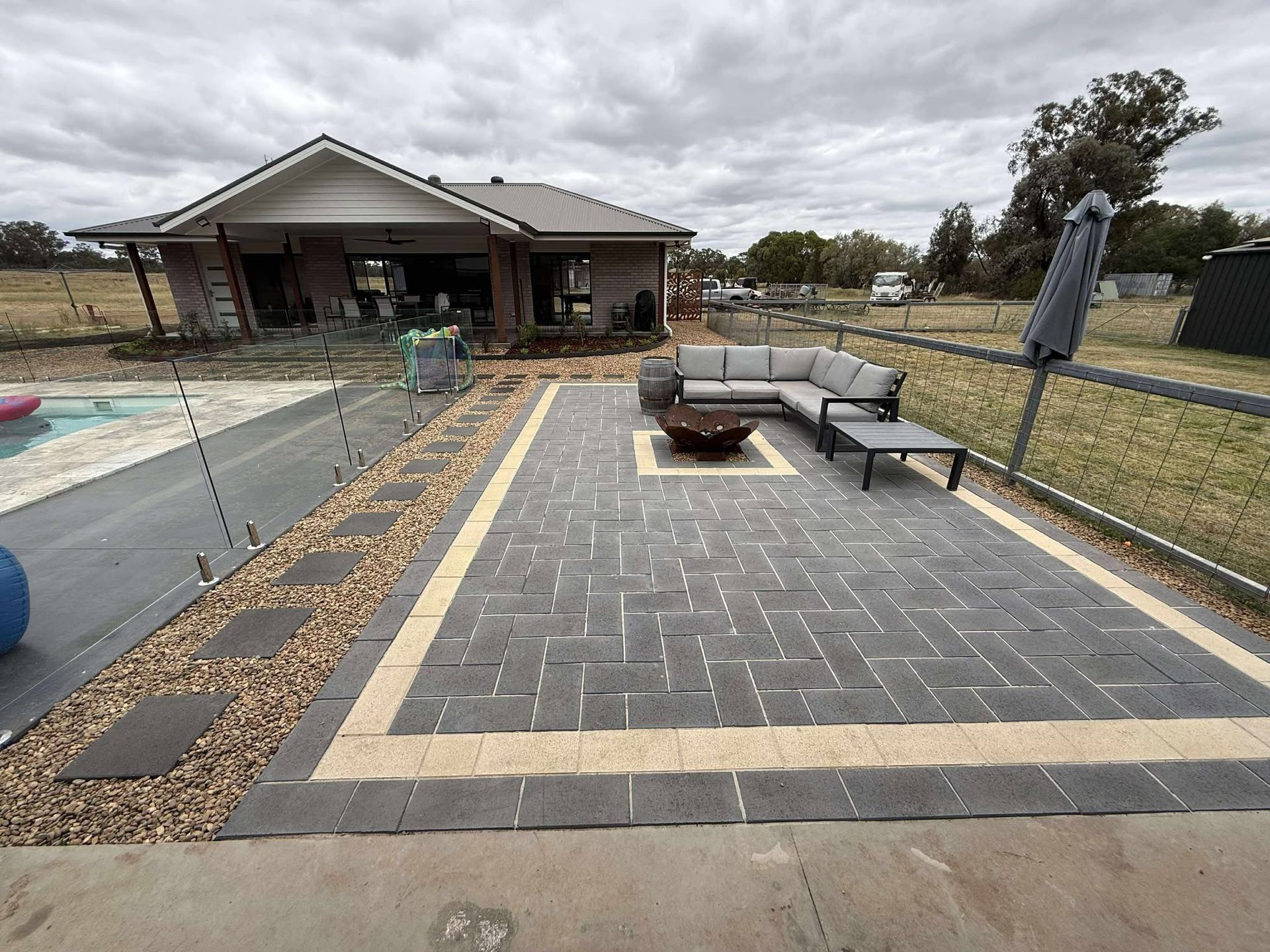 Backyard patio with dark pavers, seating, fire pit, and pool. House and fence in background — Darlington Landscaping in Dubbo, NSW