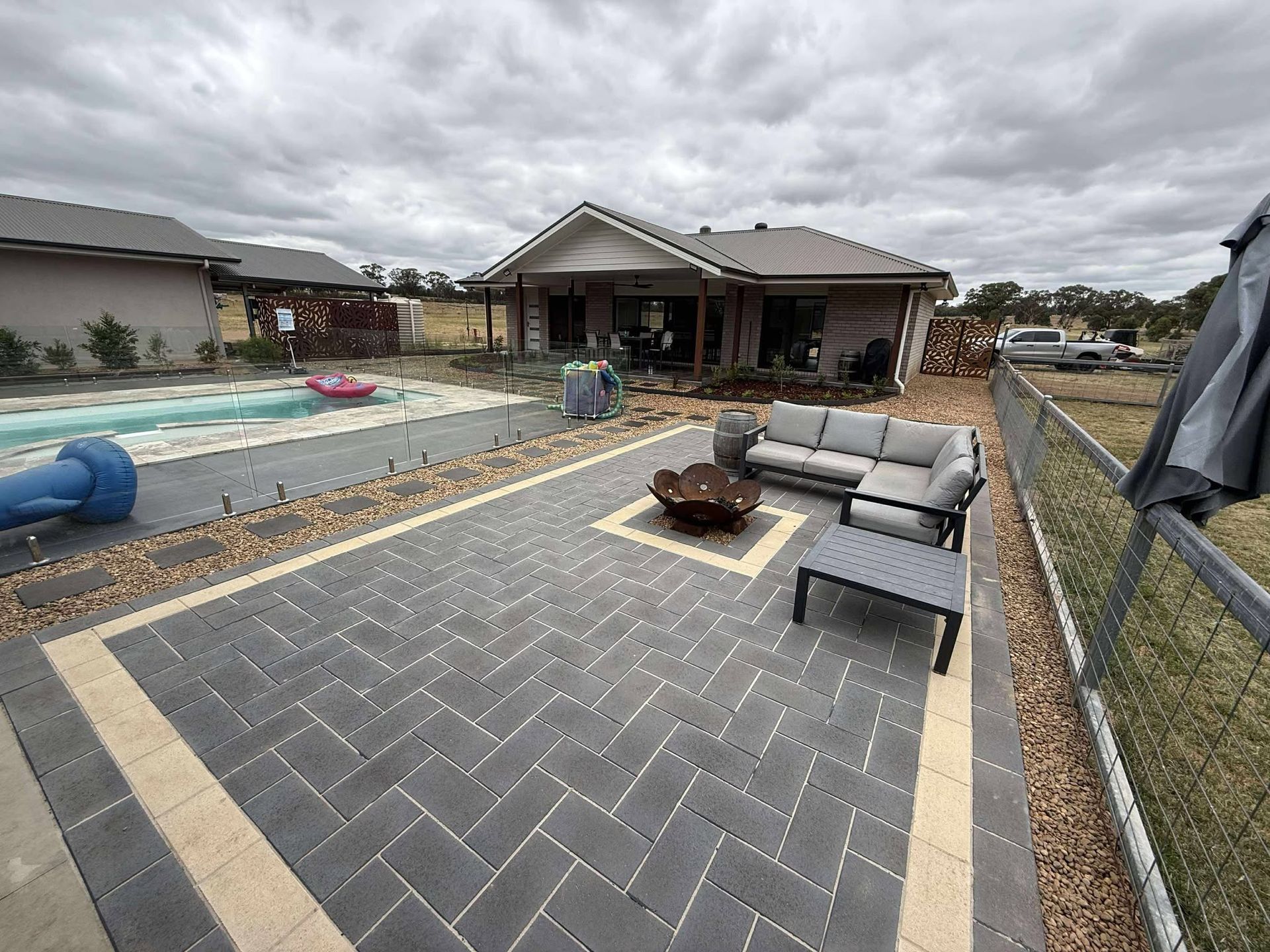 Patio with grey pavers, outdoor furniture, and a fire pit next to a house and pool under a cloudy sky — Darlington Landscaping in Mudgee, NSW