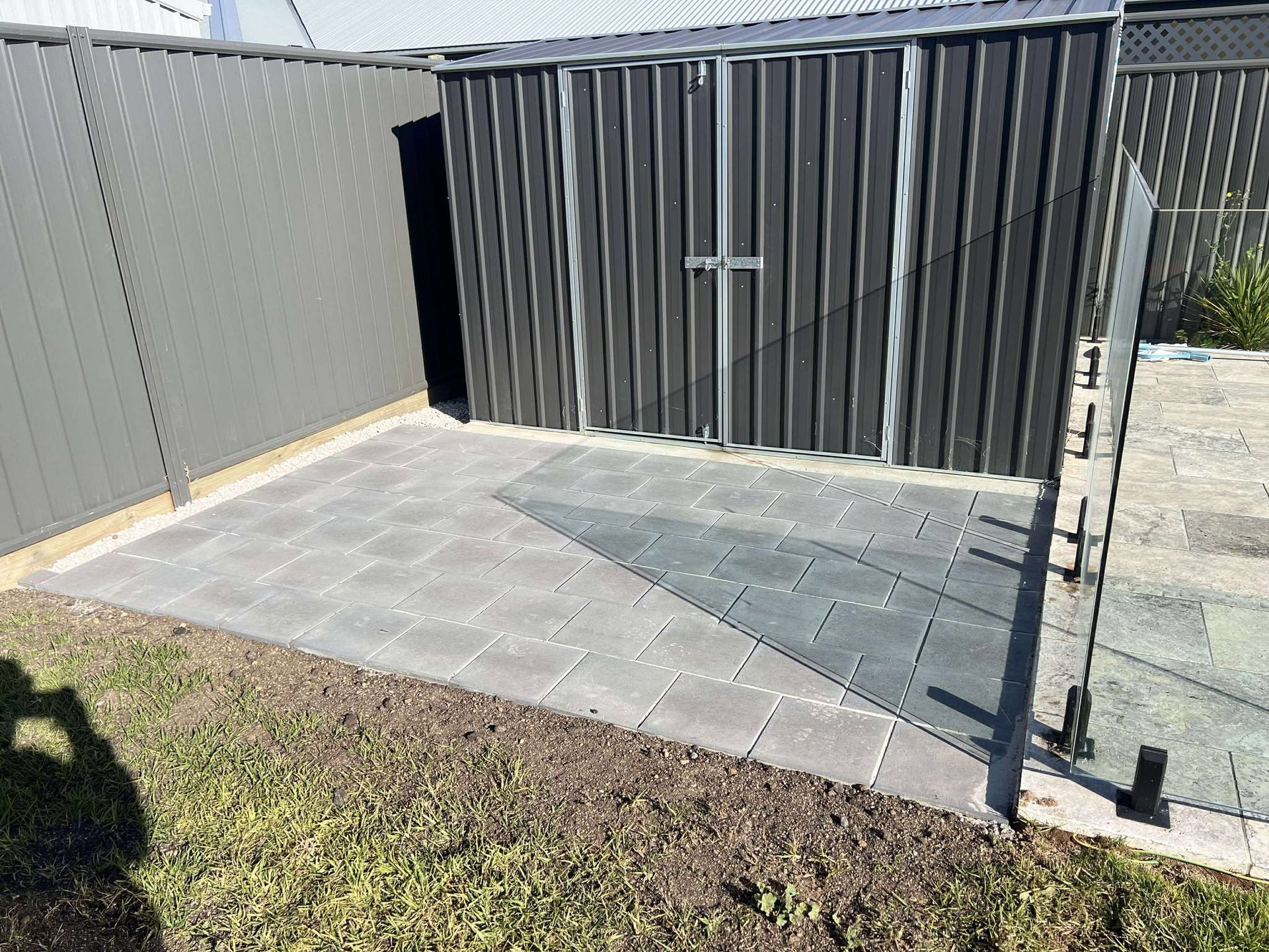Gray shed with tiled patio in a backyard, next to a fence and grass  — Darlington Landscaping in Narromine, NSW