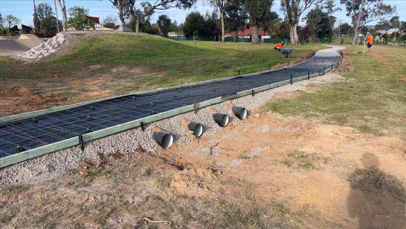 A gravel pathway with drainage pipes, surrounded by grass and dirt, with construction workers in the background — Darlington Landscaping in Wellington, NSW