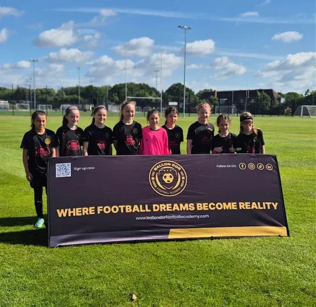 Youth soccer team posing behind a banner on a sunny field that reads “Where Football Dreams Become Reality.”