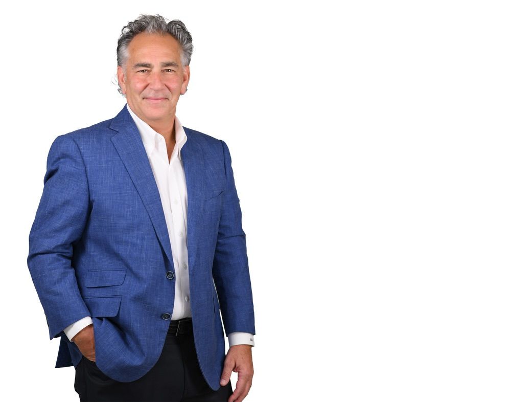 Man in blue blazer, white shirt, and dark pants, smiling with hand in pocket, against white background.