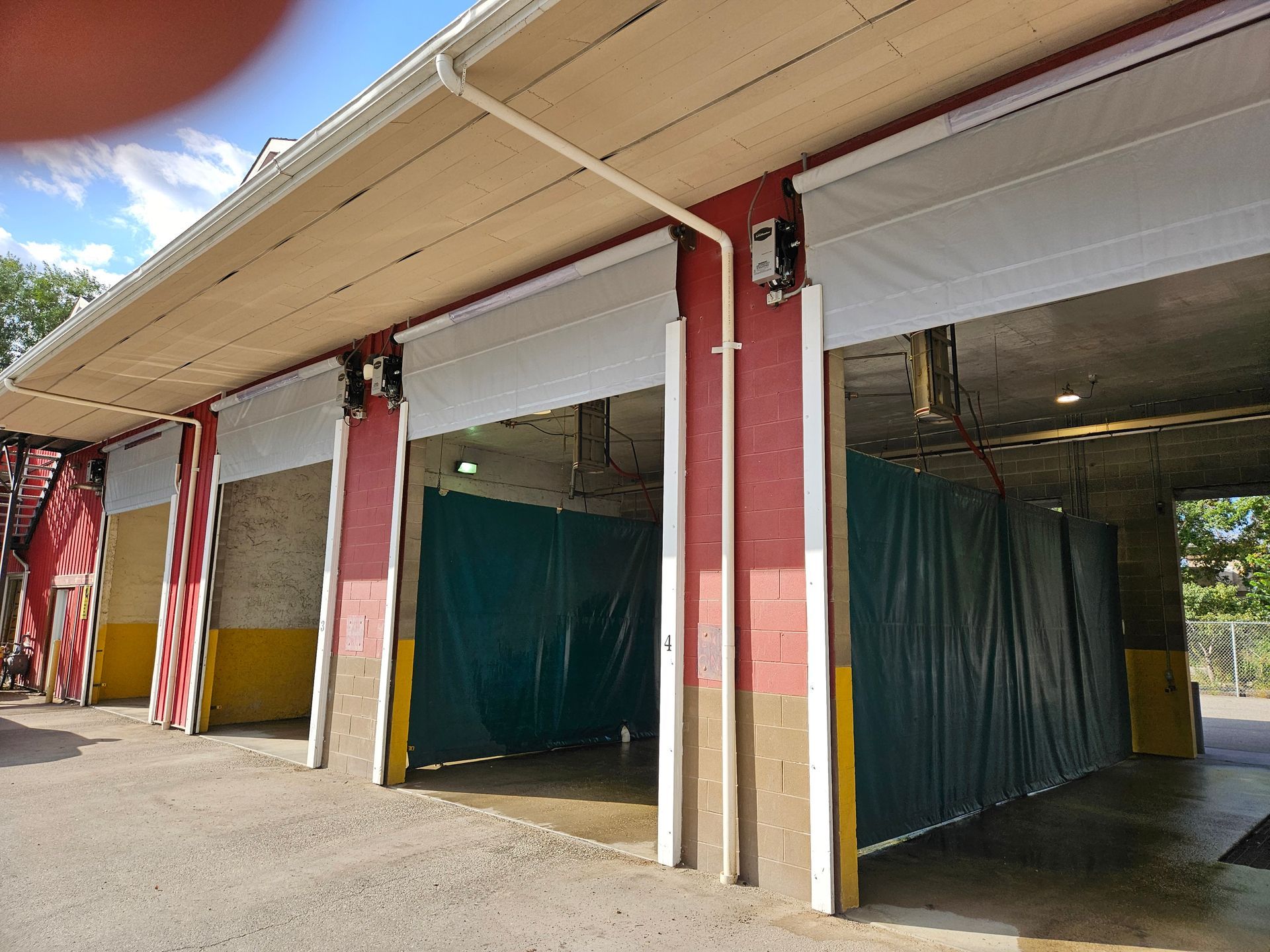 Exterior view of a car wash with open bays, roll-up doors, and beige awnings. Green curtains hang inside.