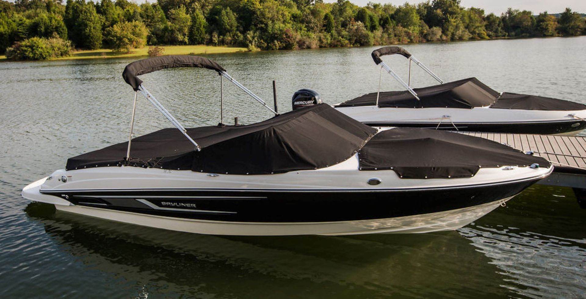 Two black and white motorboats docked on a lake, covered with black boat covers.