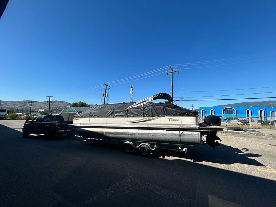 A pontoon boat on a trailer, covered with a black tarp, is towed by a black pickup truck on a sunny day.