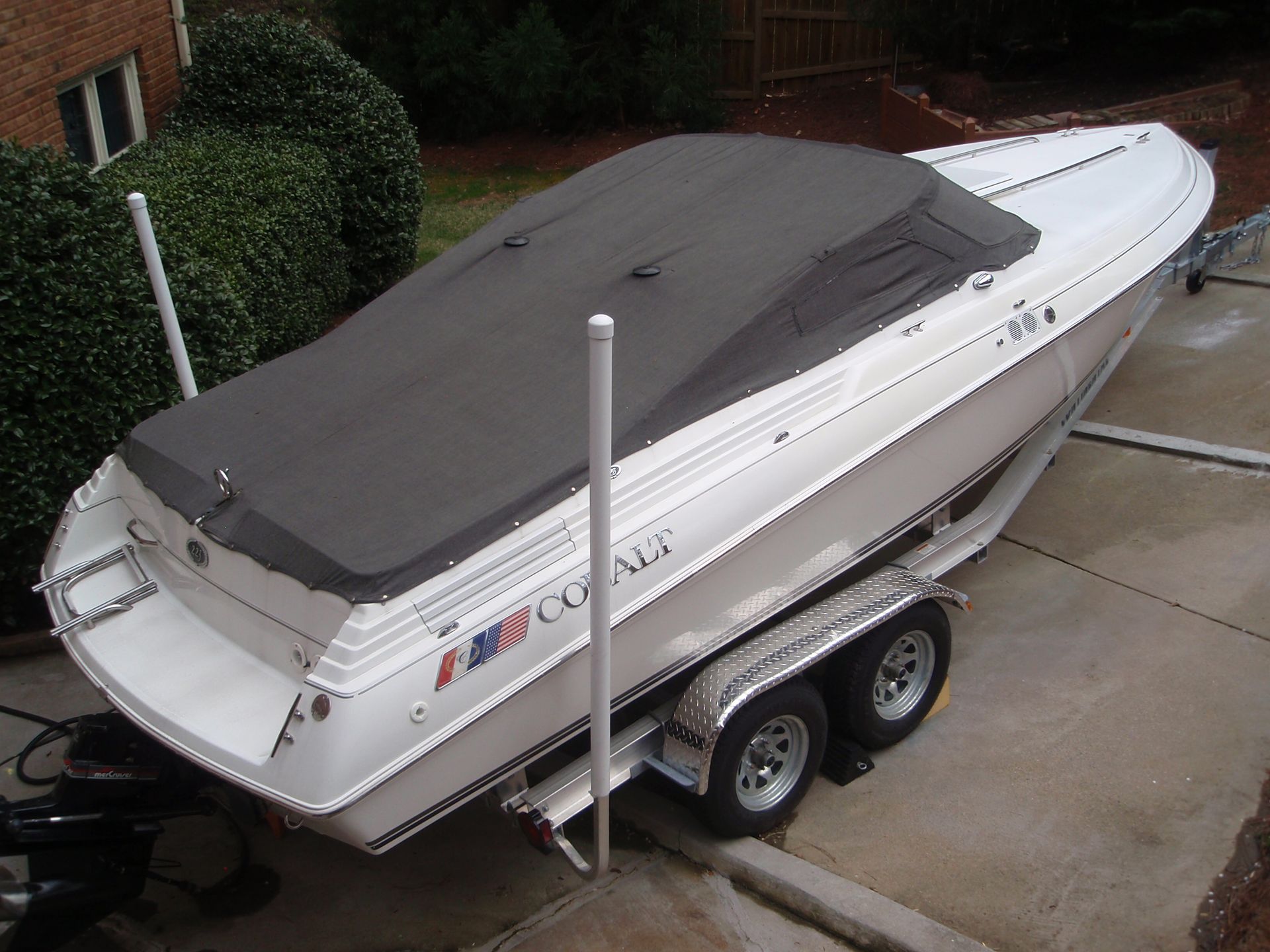 White boat on a trailer, covered with a black tarp, parked on a paved driveway.
