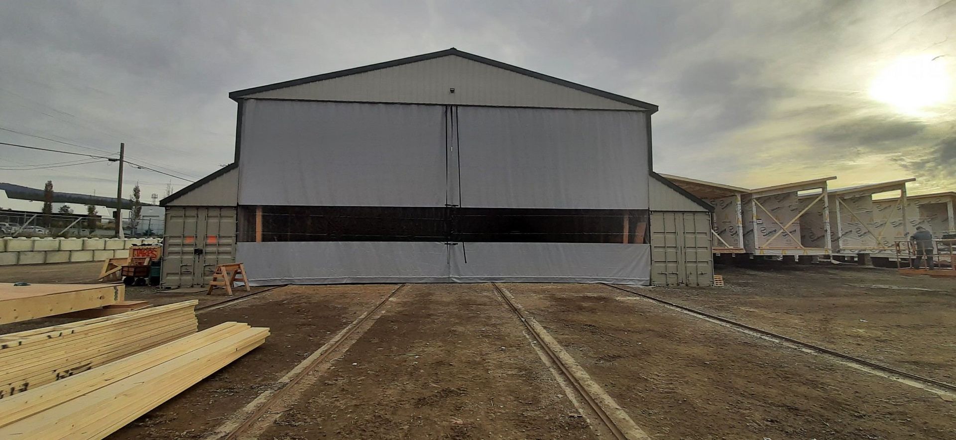 A large industrial building with a partially open door, and materials and equipment outside. Cloudy day.