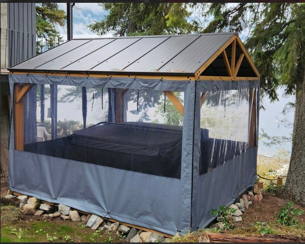 Gazebo with a black roof and gray curtains enclosing a hot tub. It's set outdoors near water.