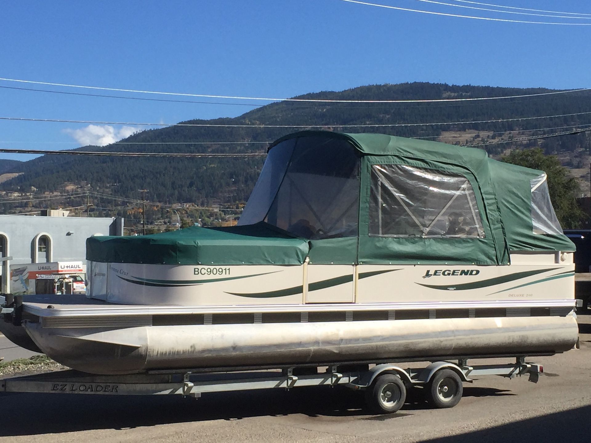 Pontoon boat on trailer, tan and green, with canvas top, in front of mountain range.