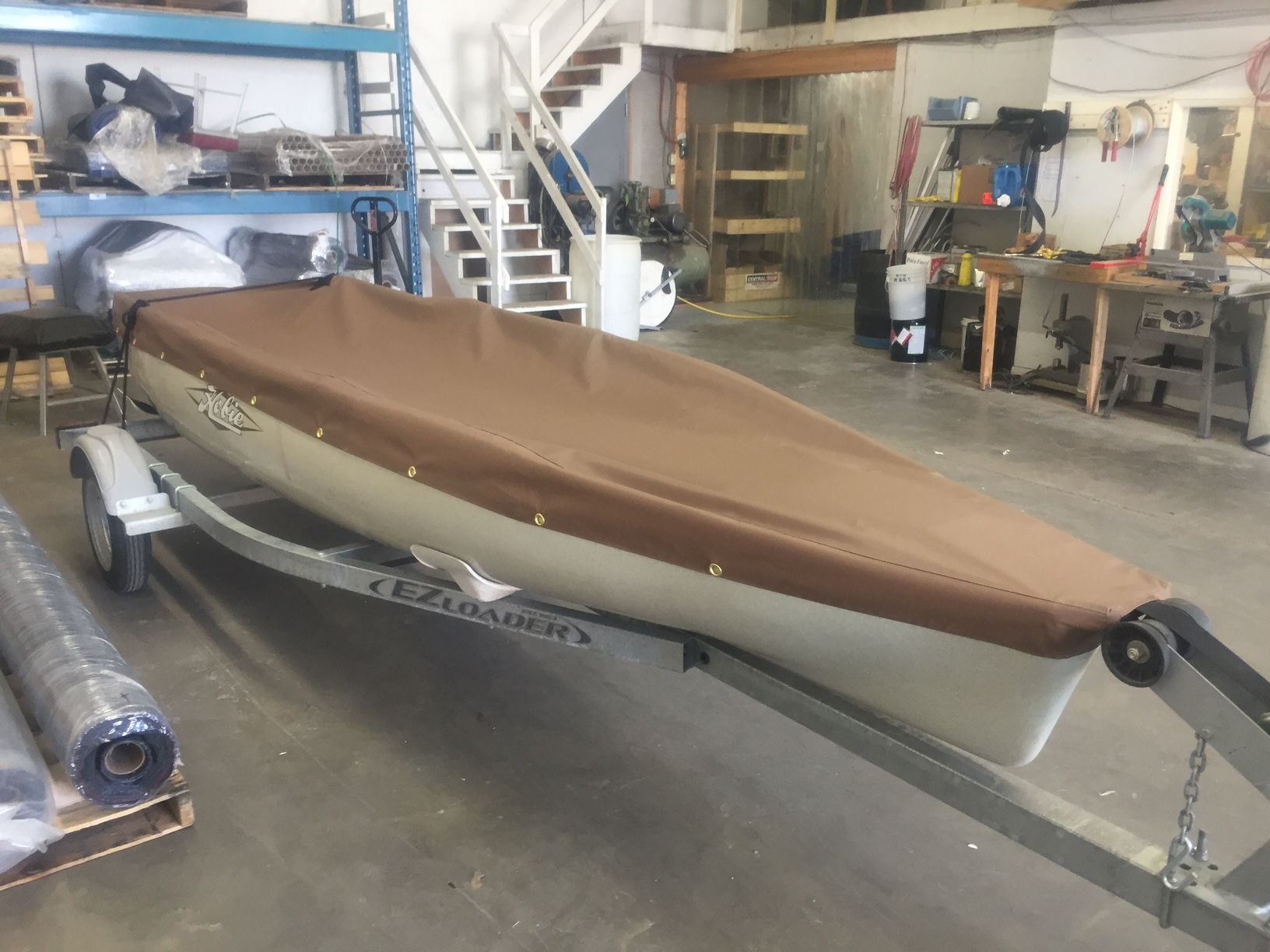 Brown-covered boat on a trailer inside a workshop, near stairs and tools.